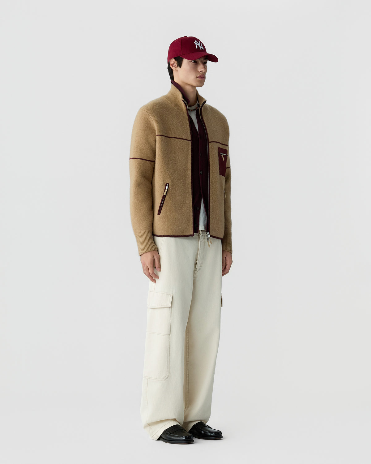 A man wearing a beige zip-up sweater with dark trim over a maroon jacket, white cargo pants, black shoes, and a red baseball cap standing against a plain white background.