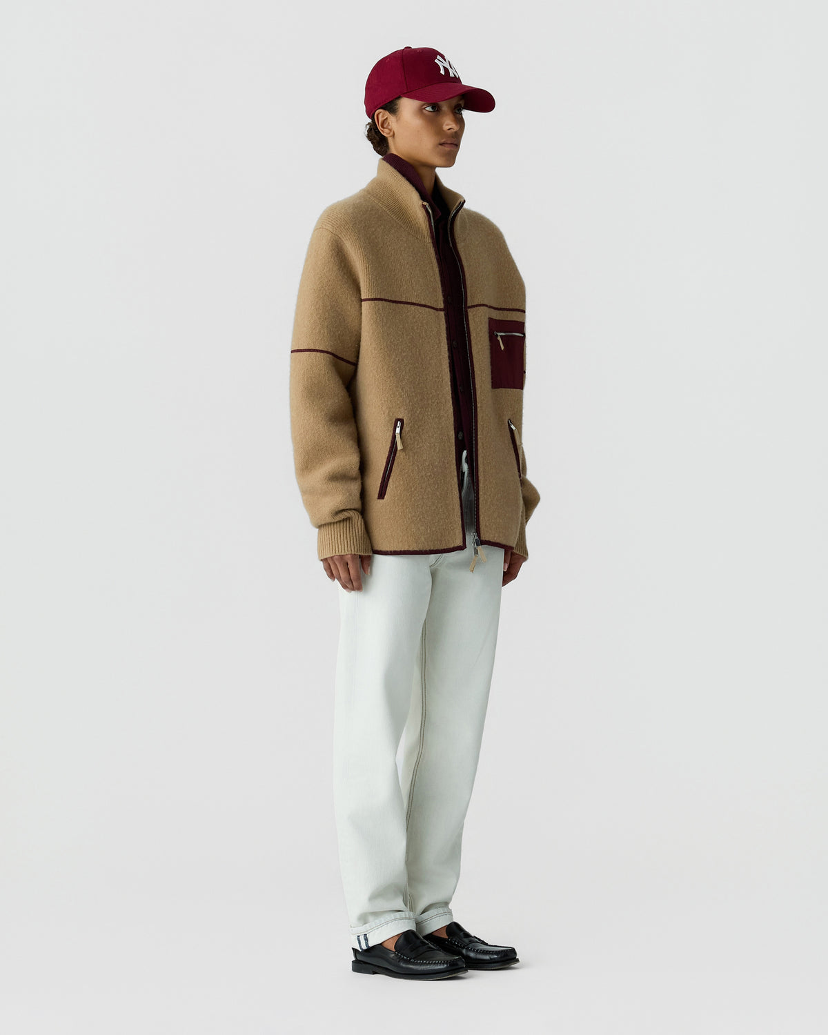 Person wearing a tan zip jacket, burgundy baseball cap, white trousers, and black loafers against a plain light gray studio background