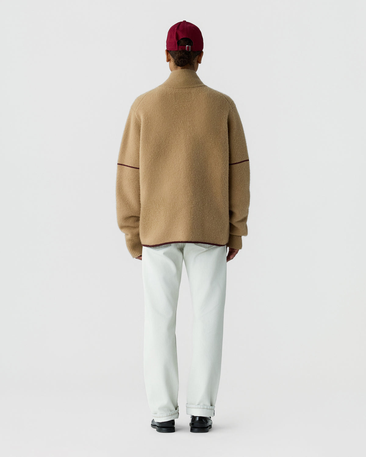 Person viewed from behind wearing a tan sweater, white trousers, burgundy cap, and black shoes against a light gray background