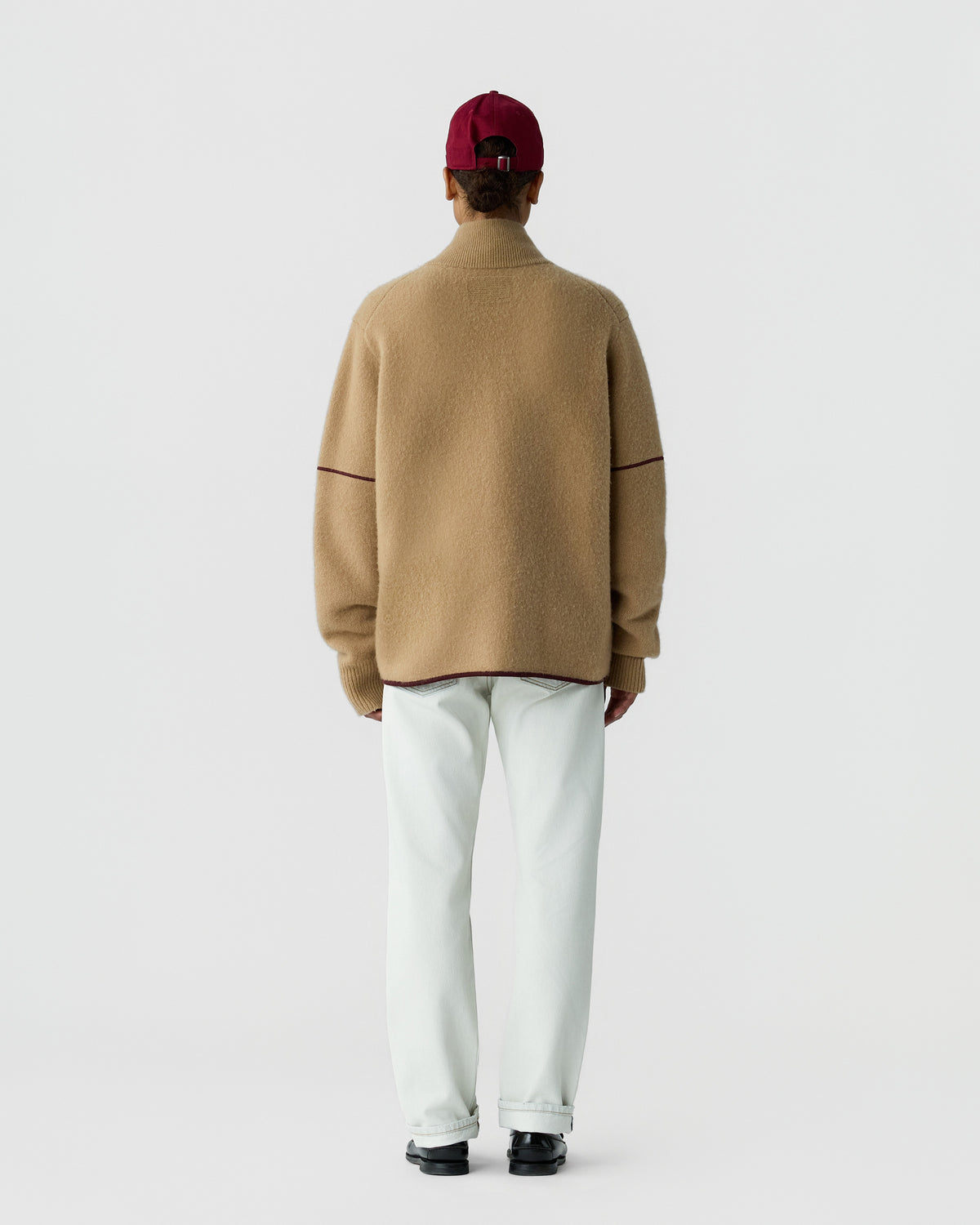 Person wearing a beige high-neck sweater with burgundy trim details, white pants, black shoes, and a red baseball cap, standing facing away against a plain background.