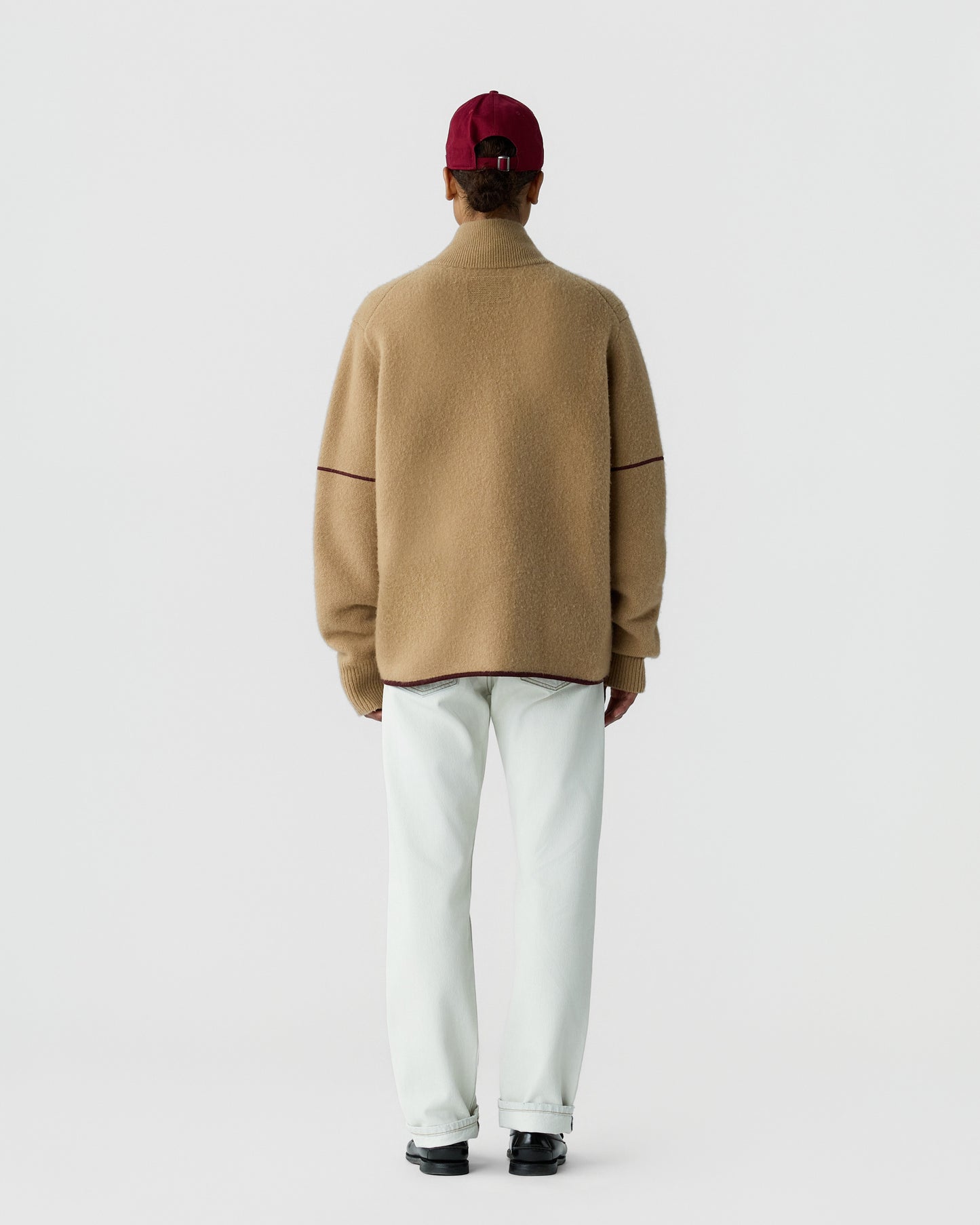Person wearing a beige high-neck sweater with burgundy trim details, white pants, black shoes, and a red baseball cap, standing facing away against a plain background.