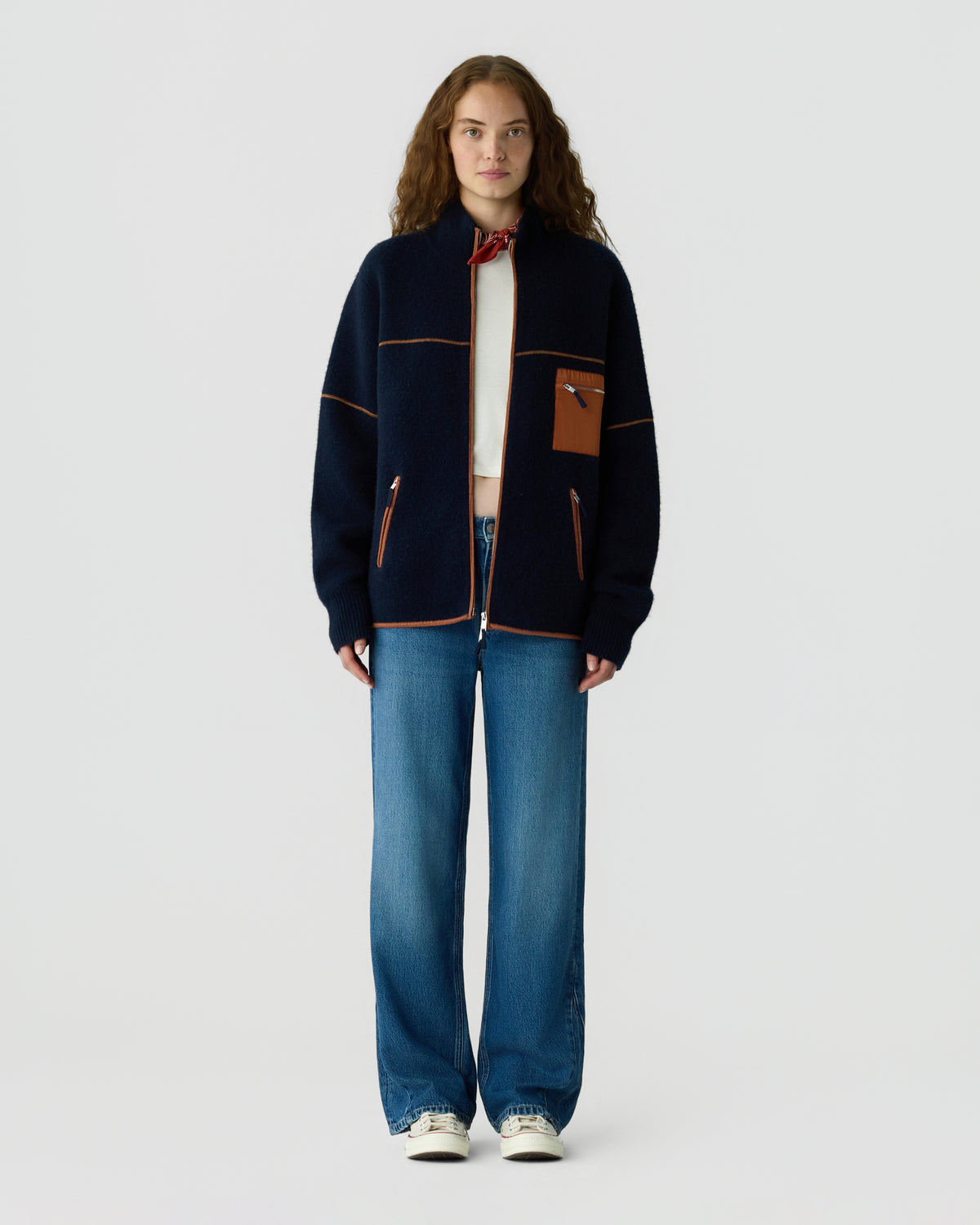 Full length portrait of a woman wearing a navy zip jacket over a white top, wide leg blue jeans, and white sneakers against a light gray background