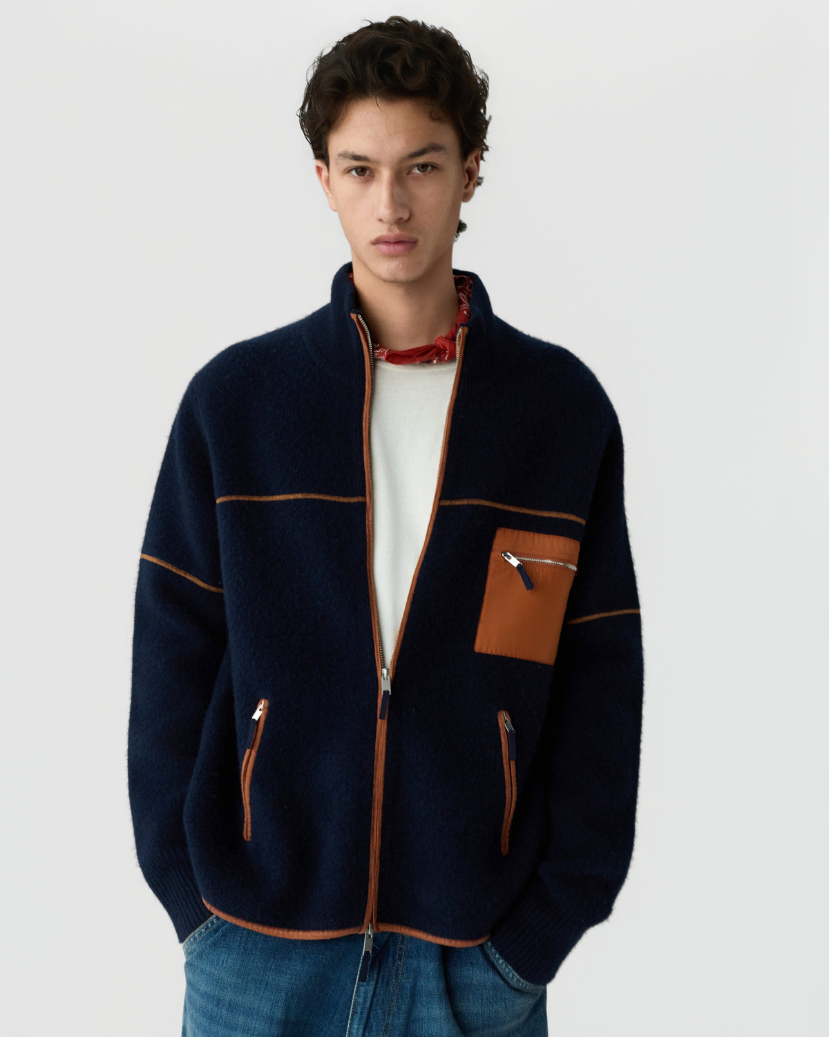Young man wearing a navy blue zip-up fleece jacket with brown trim and zippered pockets over a white shirt and blue jeans against a plain background.