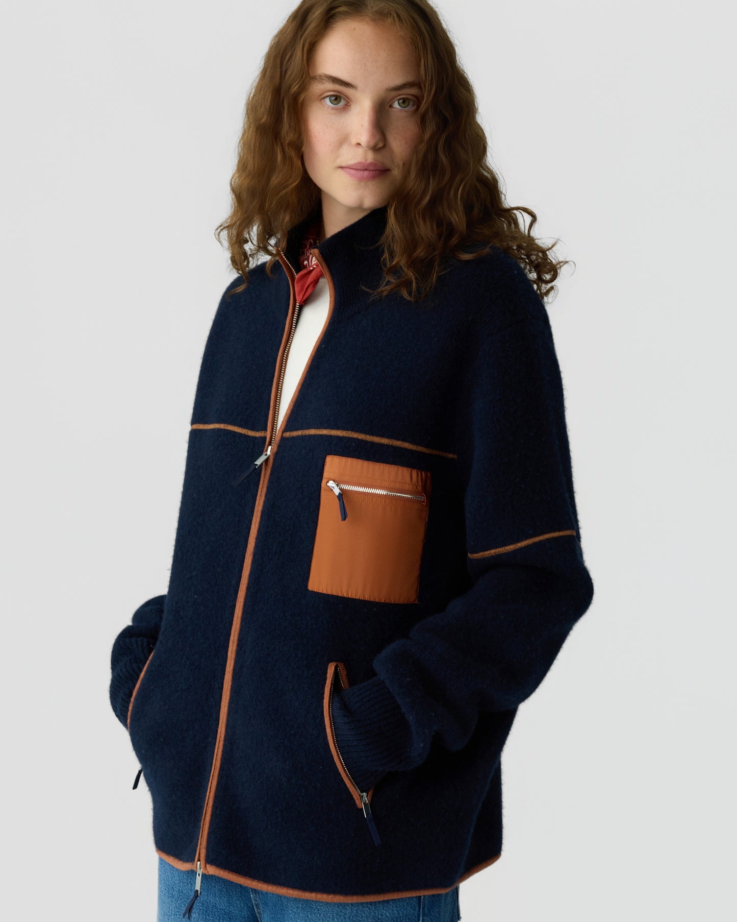 Woman wearing a navy blue zip-up jacket with rust-colored trim and a chest pocket, standing with hands in pockets against a plain light background.