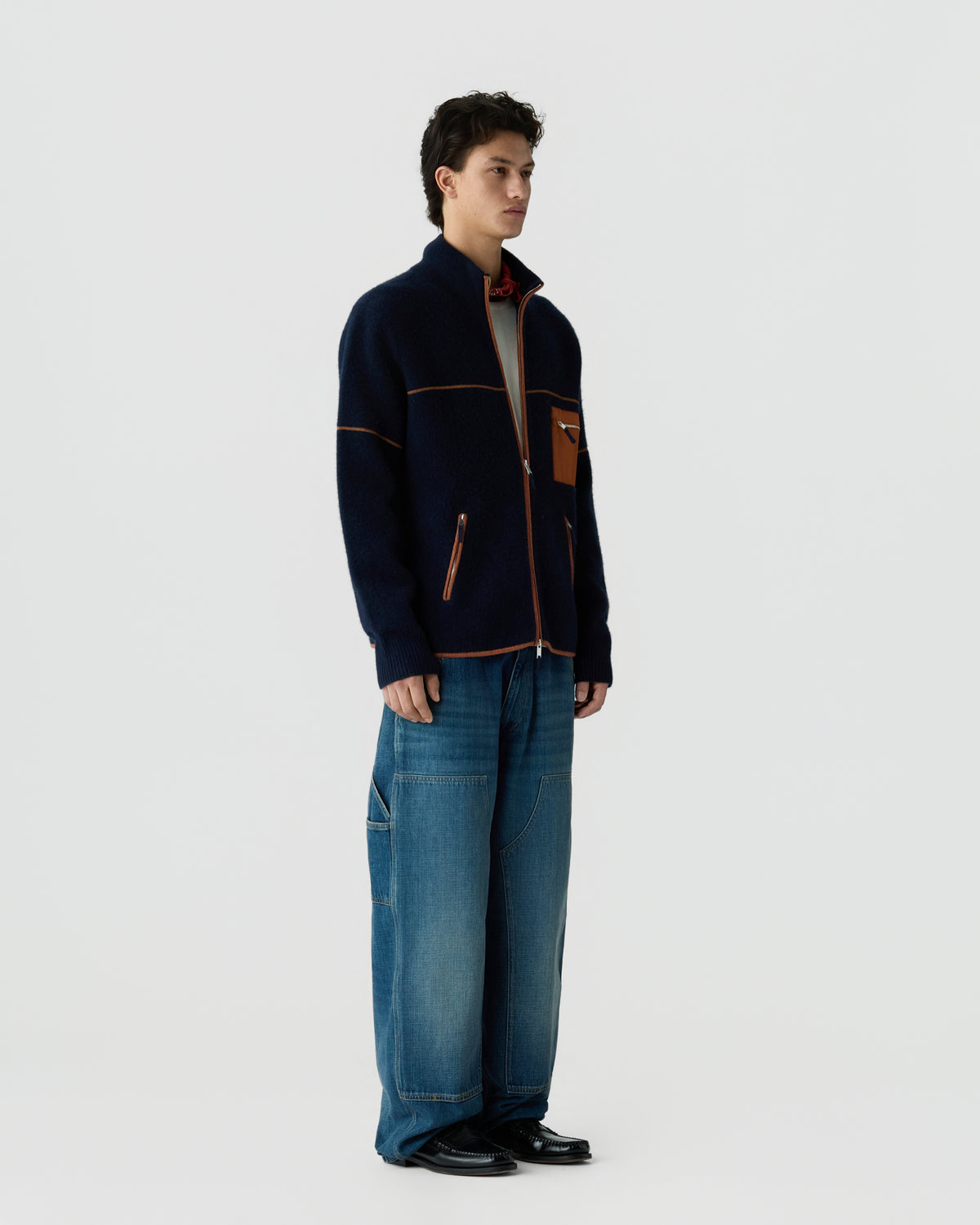 Young man wearing a navy zip-up cardigan with brown pocket details and wide-leg blue jeans, standing against a plain light gray background.