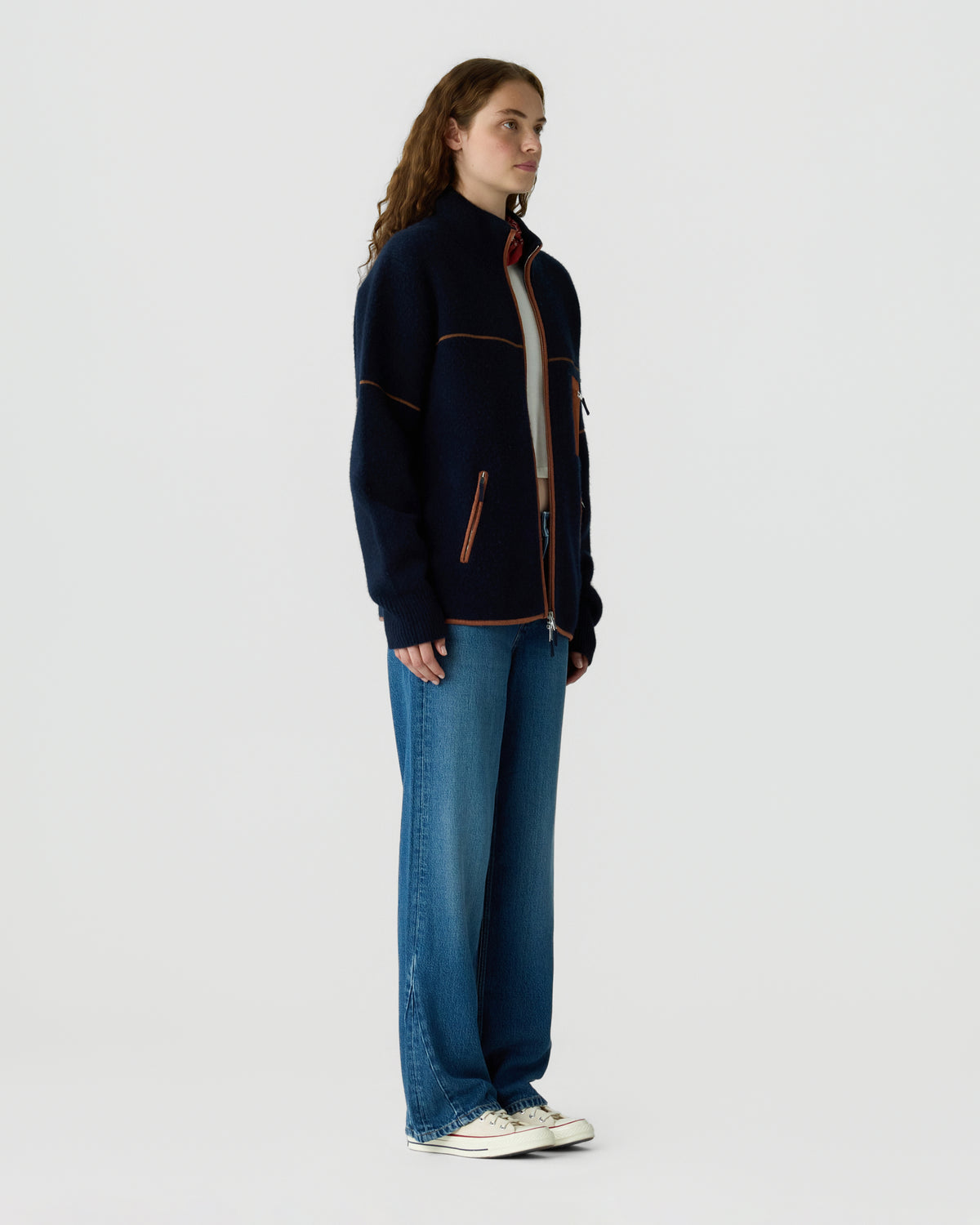 Full length view of a woman wearing a navy zip jacket with orange trim blue jeans and white sneakers against a light gray background