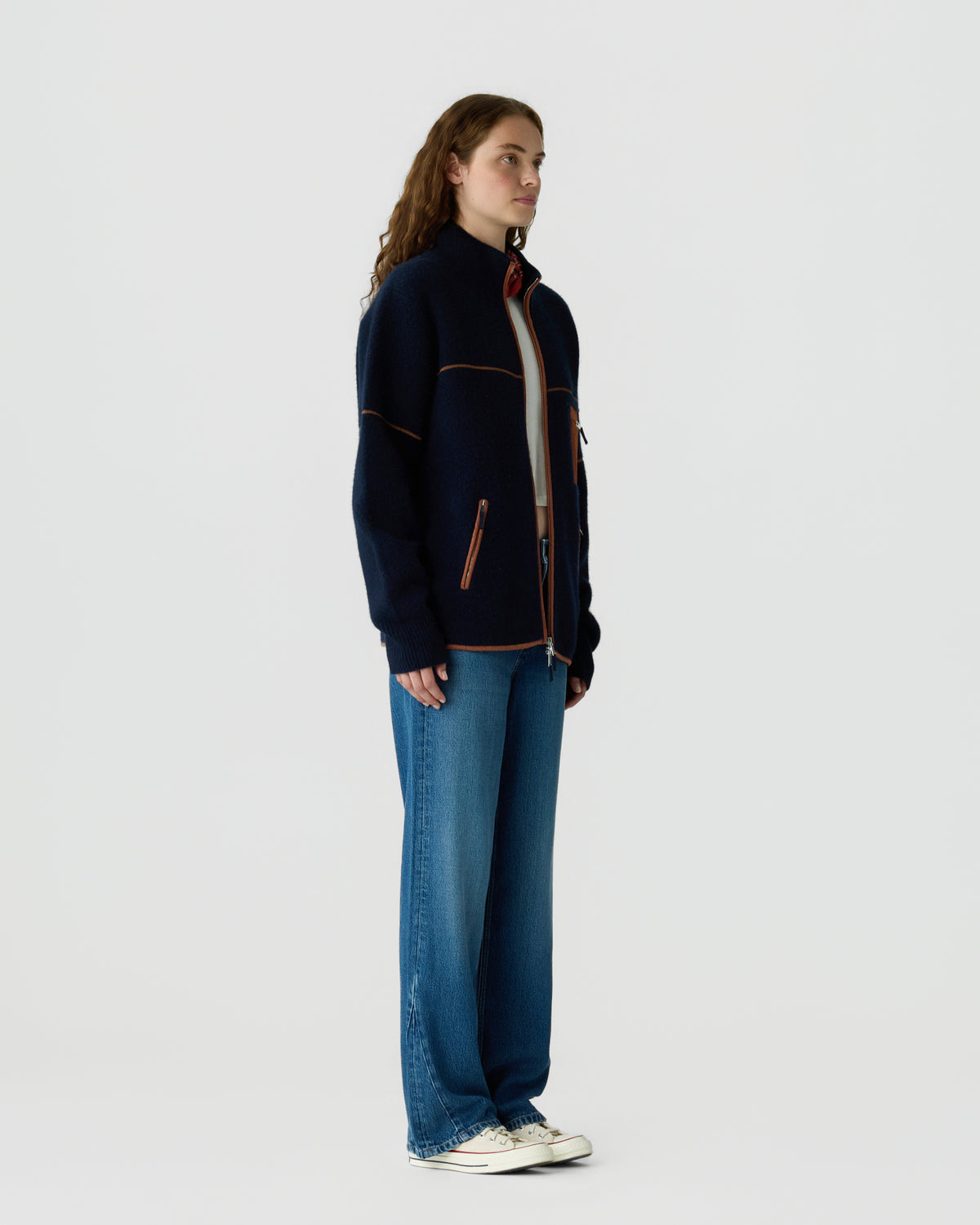 Side view of a woman with long curly hair wearing a navy blue zip-up jacket with orange piping, wide-leg blue jeans, and white sneakers against a plain background.