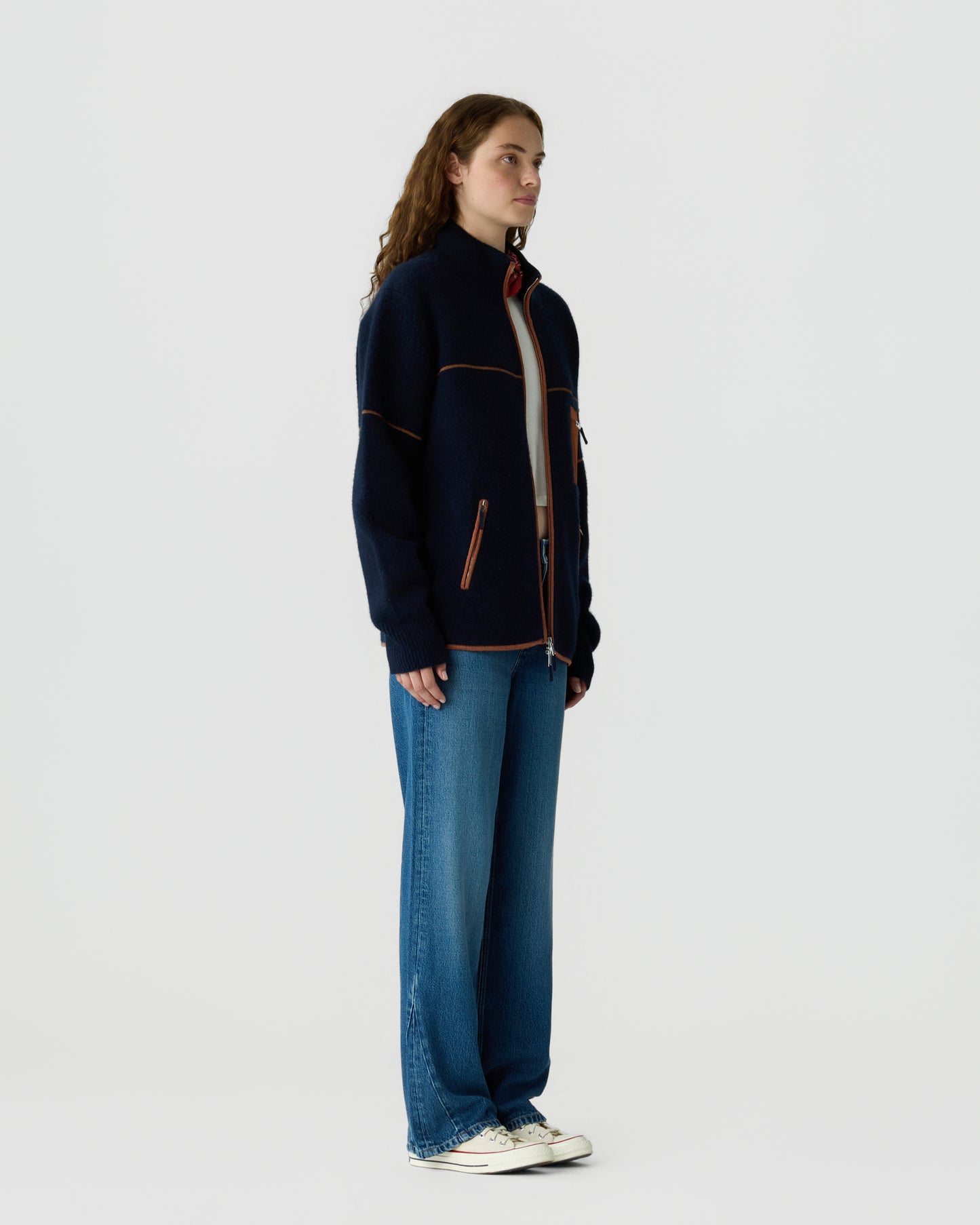 Side view of a woman with long curly hair wearing a navy blue zip-up jacket with orange piping, wide-leg blue jeans, and white sneakers against a plain background.