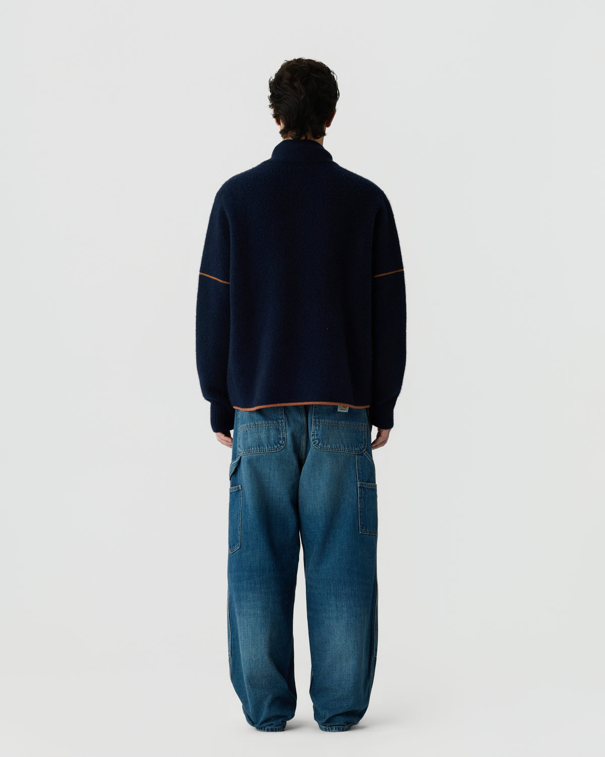 Rear view of a person wearing a dark navy cashmere sweater with contrasting trim and loose-fitting blue denim jeans against a plain gray background.