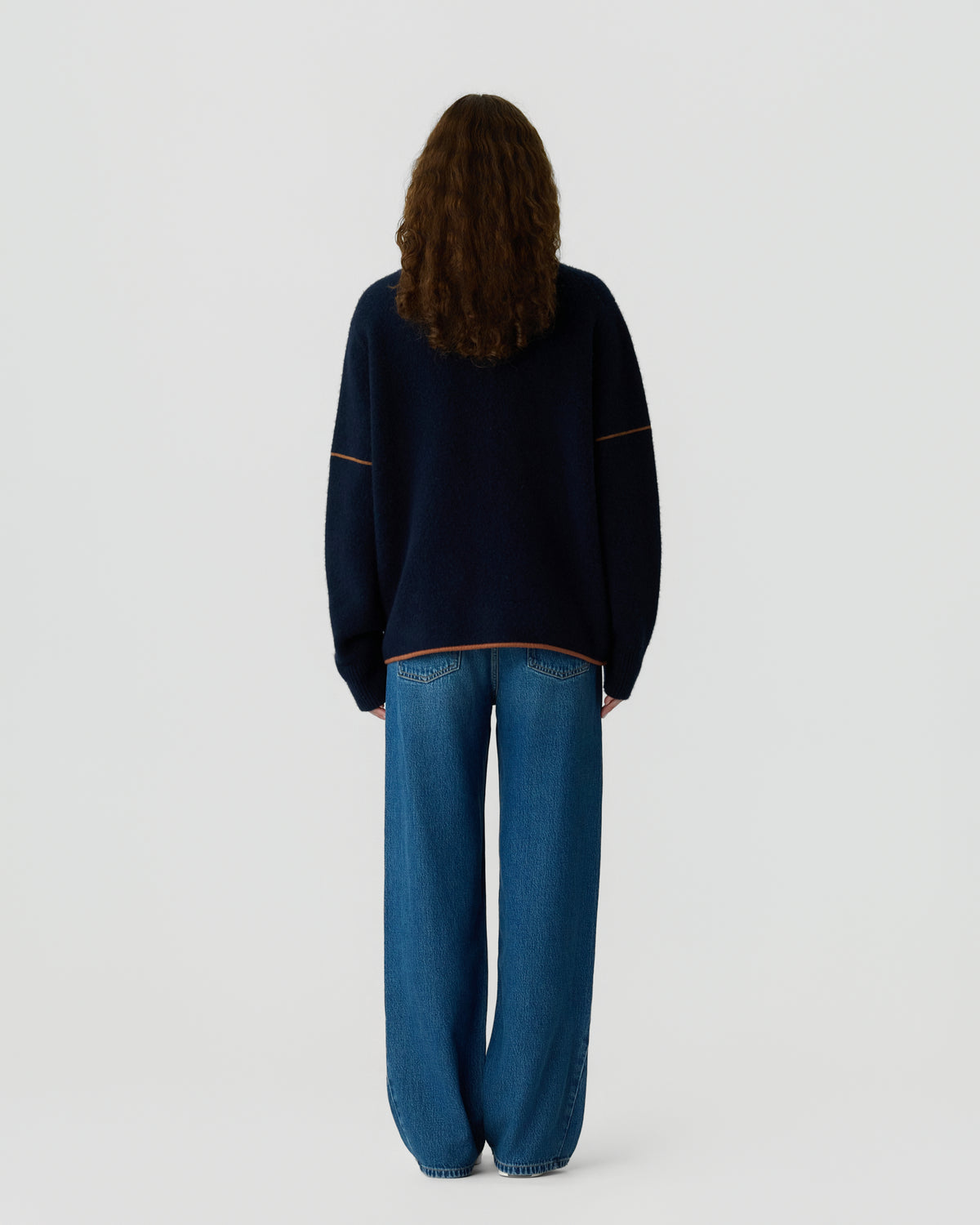 Person with long curly hair seen from behind wearing a loose dark navy sweater with light stitching and wide-leg blue jeans against a white background.