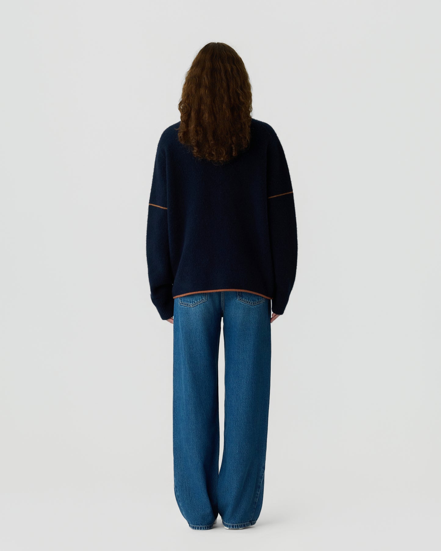 Person with long curly hair seen from behind wearing a loose dark navy sweater with light stitching and wide-leg blue jeans against a white background.