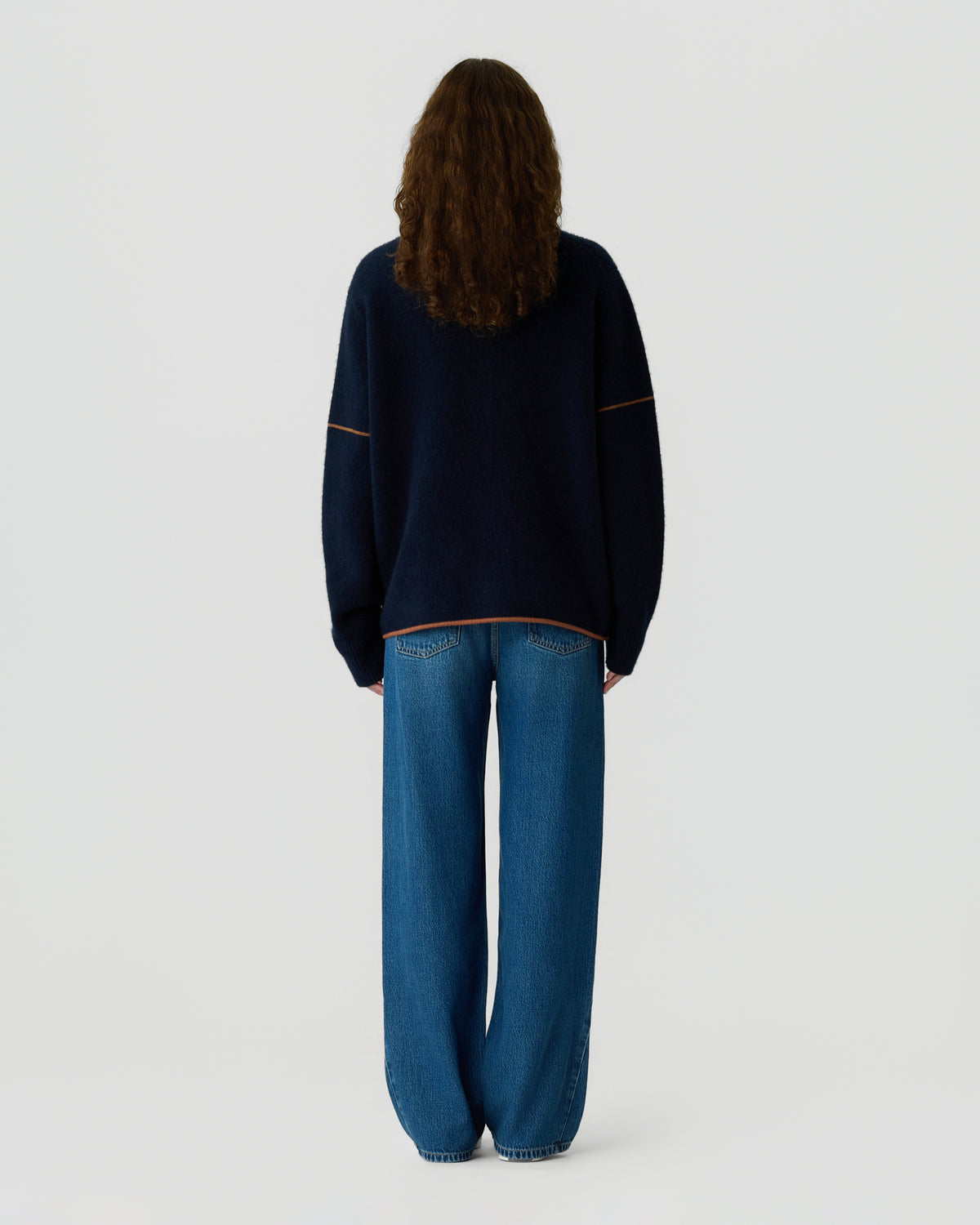 Back view of a person with long brown hair wearing a dark navy sweater with orange trim and relaxed blue jeans against a light gray background