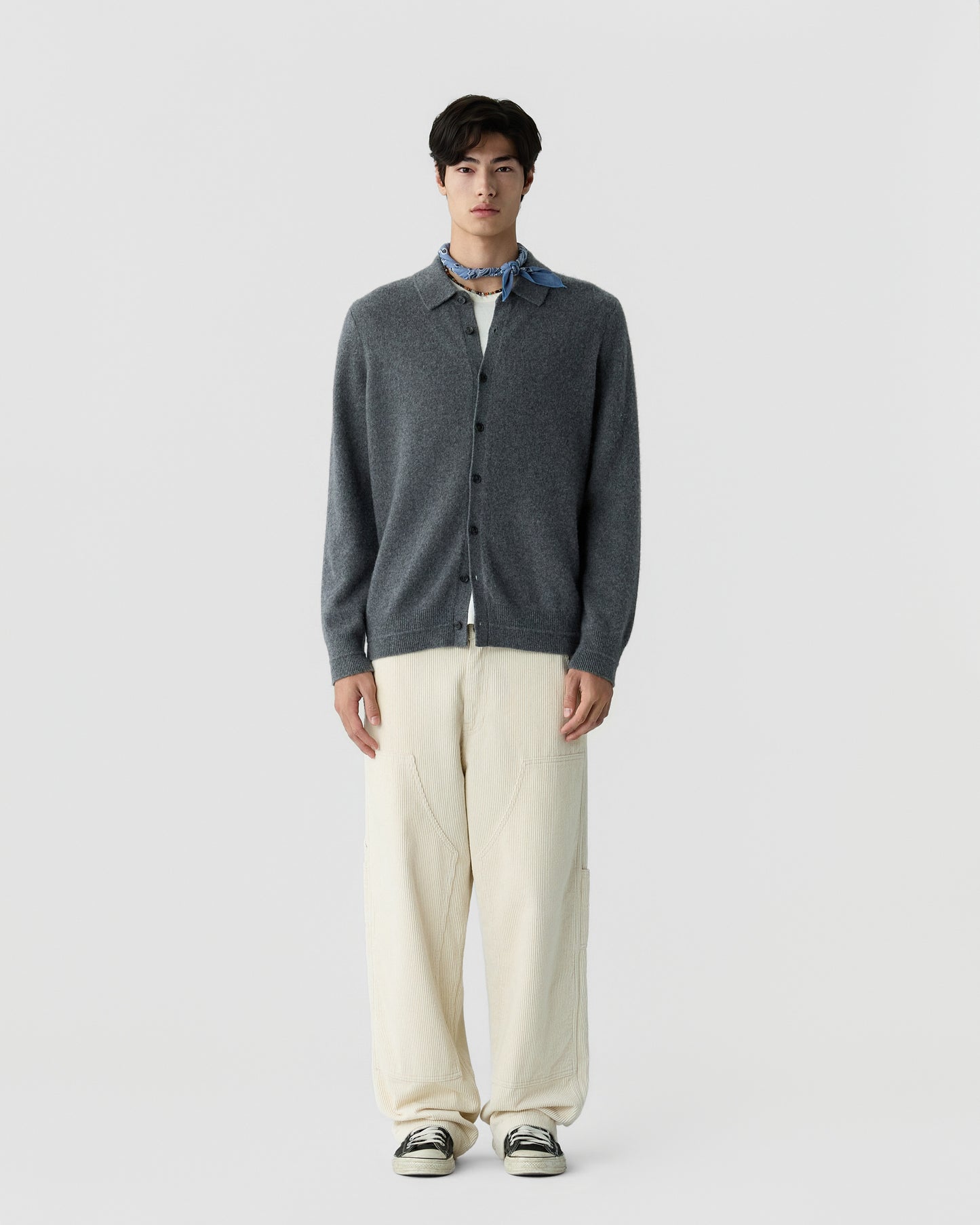 Young man wearing a dark gray buttoned cardigan over a white shirt, cream wide-legged pants, black and white sneakers, and a blue scarf tied around his neck, standing against a plain light gray background.