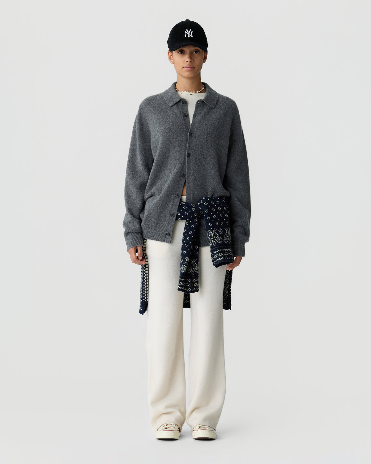 Model wearing a gray button front cardigan over a patterned navy top, white trousers, cream shoes, and a black baseball cap against a light gray background