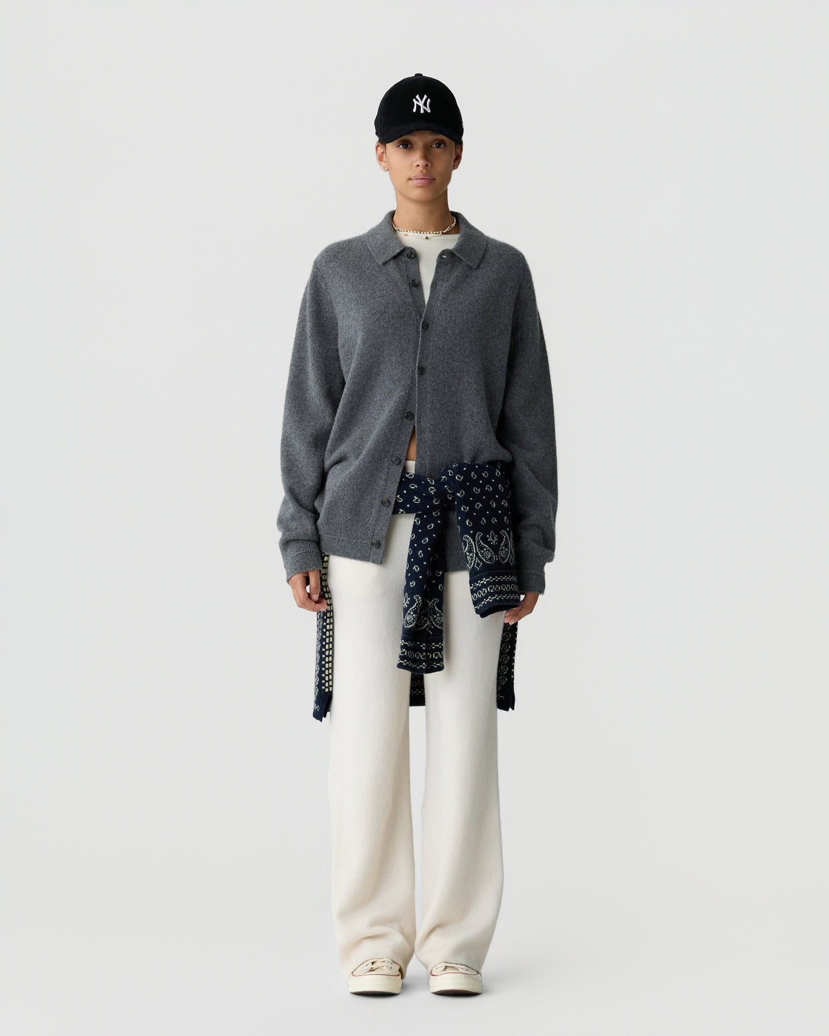 Woman wearing a dark gray button-up cashmere cardigan over a white top with wide-leg white pants, a navy patterned sweater tied at the waist, white sneakers, and a black baseball cap against a plain white background.