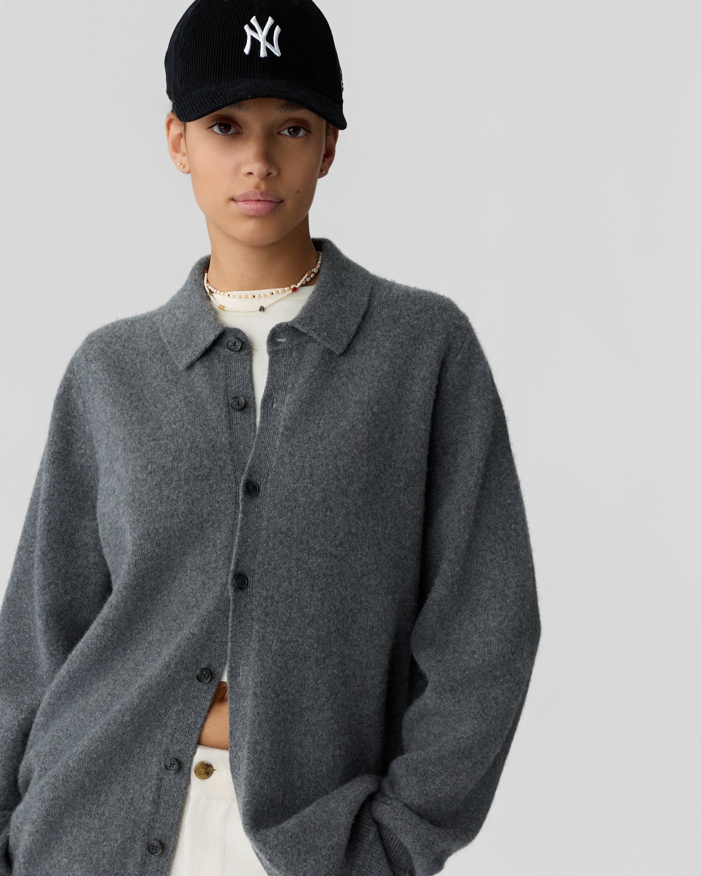 Young woman wearing a cozy grey button-up cashmere cardigan over a white top, paired with a black baseball cap featuring a white logo, standing against a plain background.