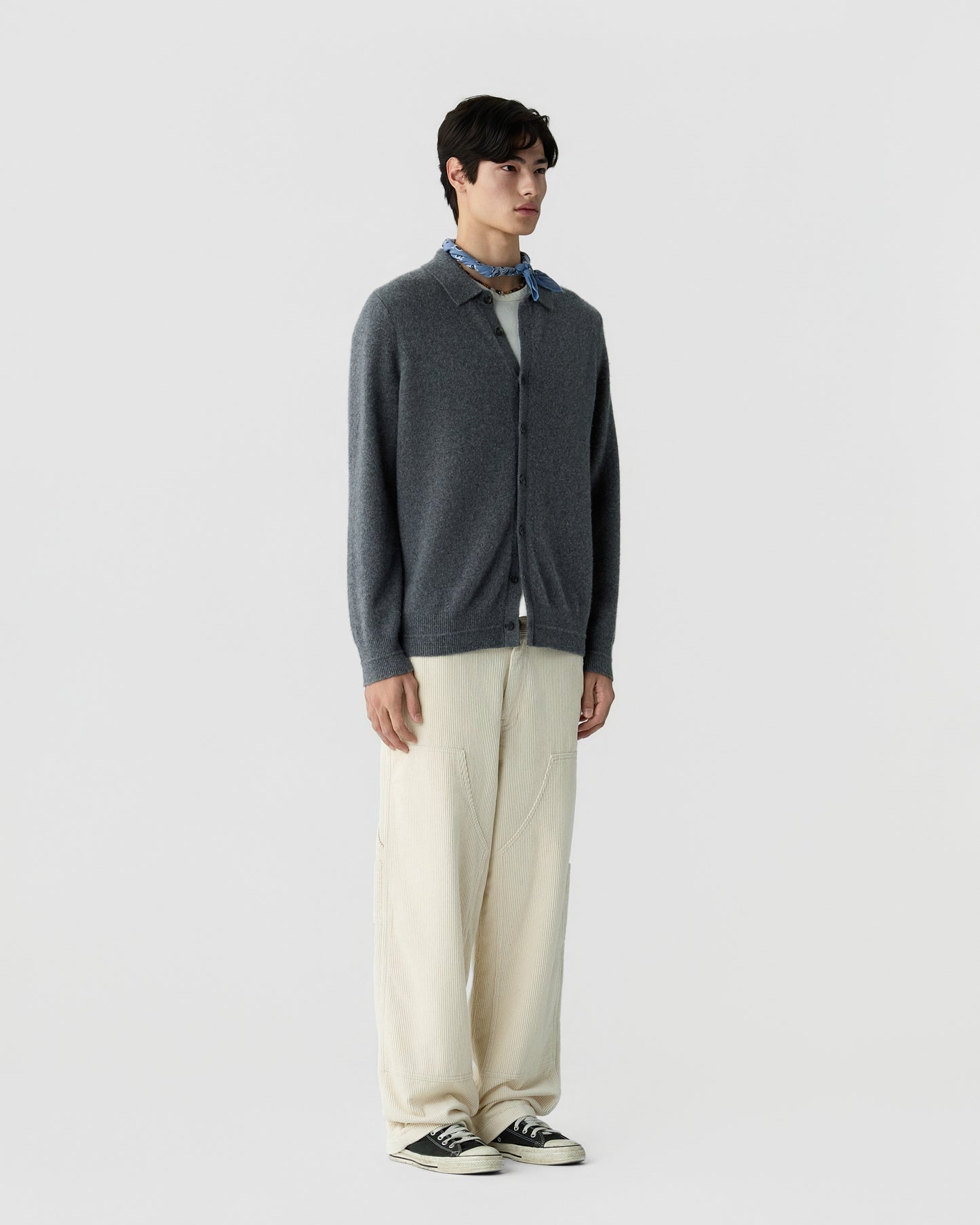 Young man wearing a dark gray button-up cashmere cardigan, light beige wide-leg pants, black and white sneakers, and a blue patterned neck scarf, standing against a plain background.
