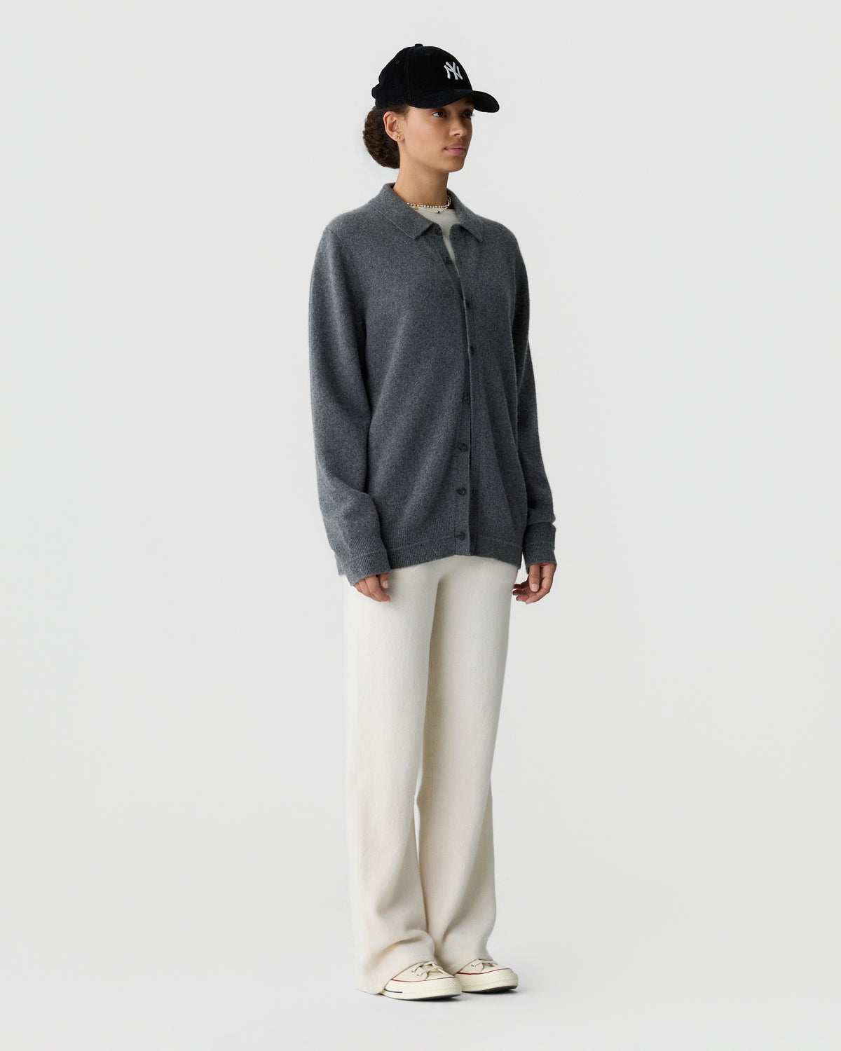 Model wearing a charcoal gray button front cardigan, cream wide leg trousers, white sneakers, and a black baseball cap against a light gray background