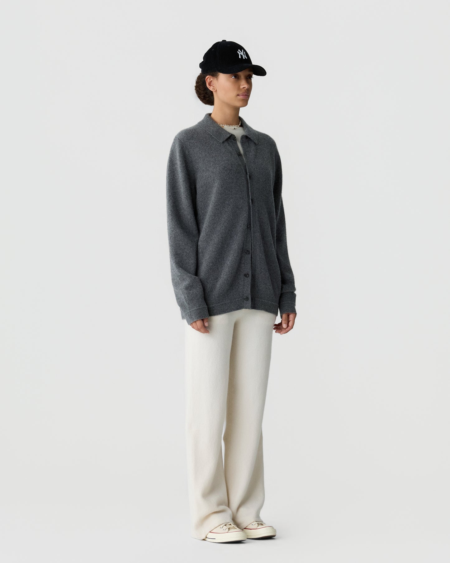 A person wearing a dark gray button-up cardigan over a light shirt, cream wide-leg pants, white sneakers, and a black baseball cap standing against a plain white background.