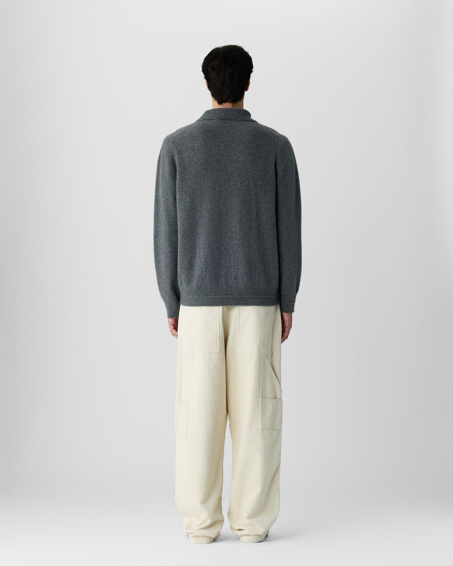 Person standing facing away wearing a dark gray long-sleeve cashmere sweater and loose-fitting cream-colored pants against a plain light gray background.