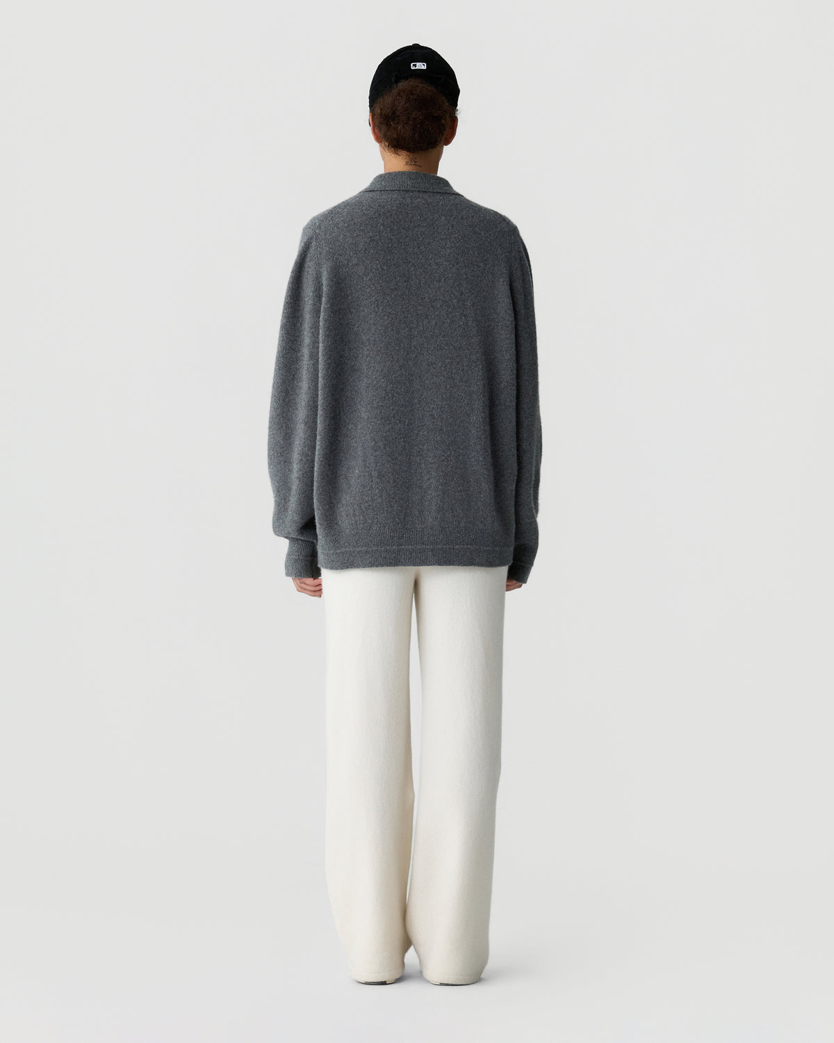 Back view of a person wearing a loose gray sweater, white trousers, and a black cap against a light gray studio background