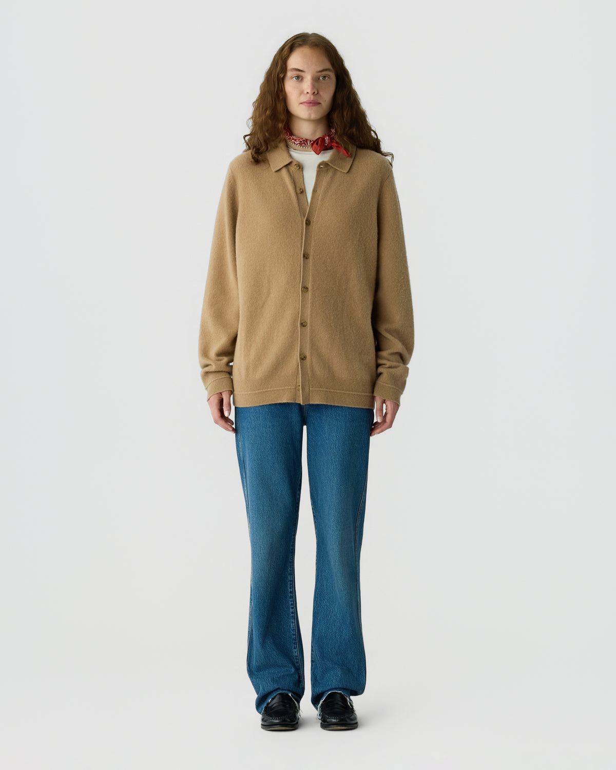 Full-length portrait of a woman wearing a camel button-front cardigan over a collared shirt with blue jeans and black shoes against a light gray background