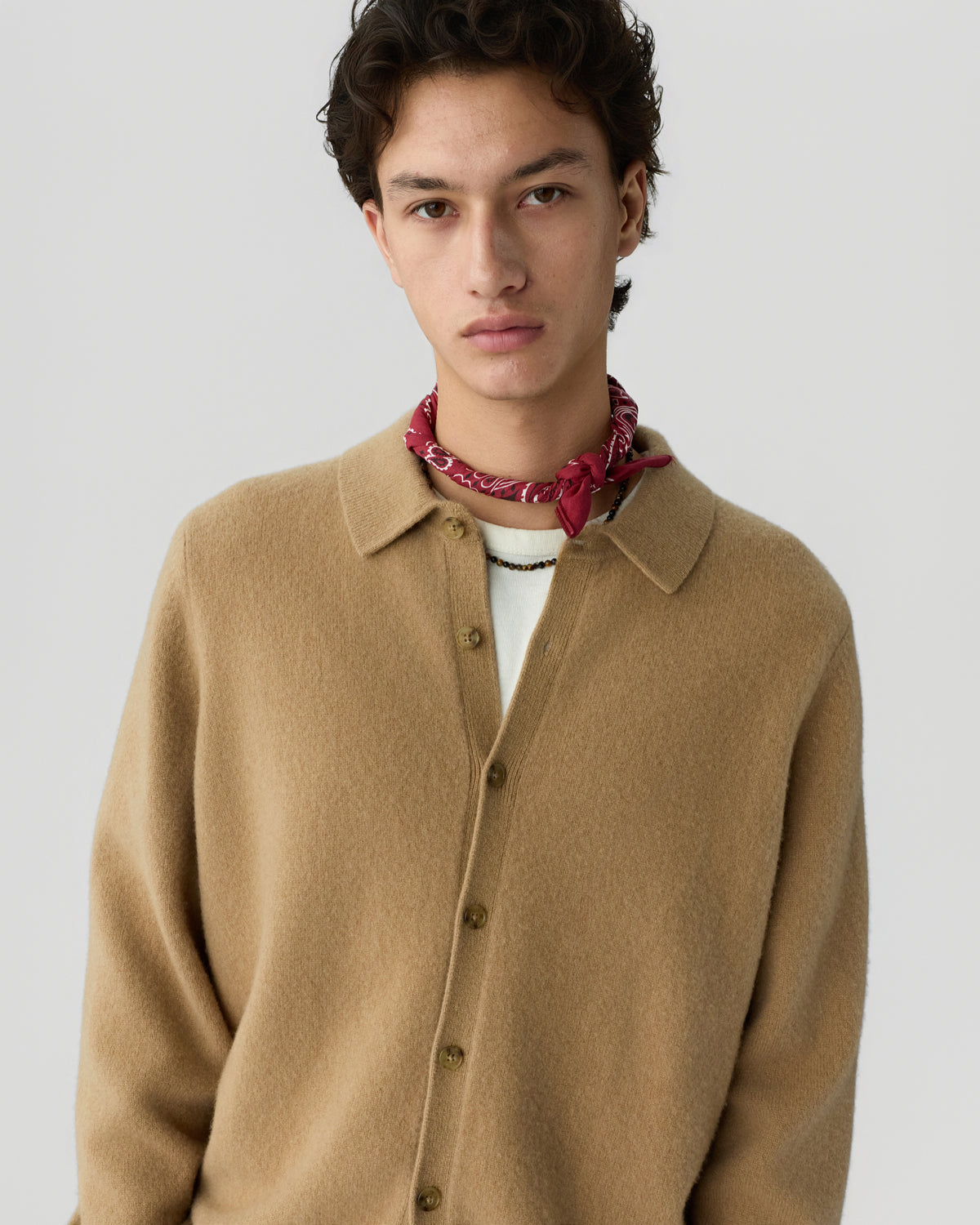 Young man wearing a camel-colored button-up cashmere shirt over a white top, accessorized with a red bandana tied around his neck against a plain light background.