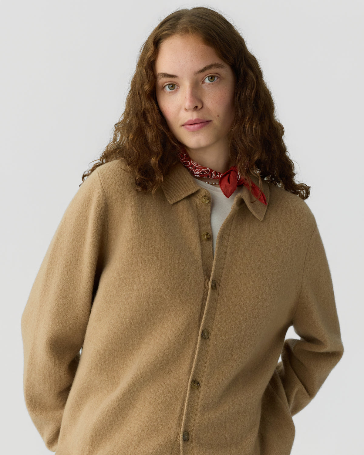 Young woman with curly brown hair wearing a camel-colored button-up cashmere sweater and a red patterned neck scarf against a plain light background.