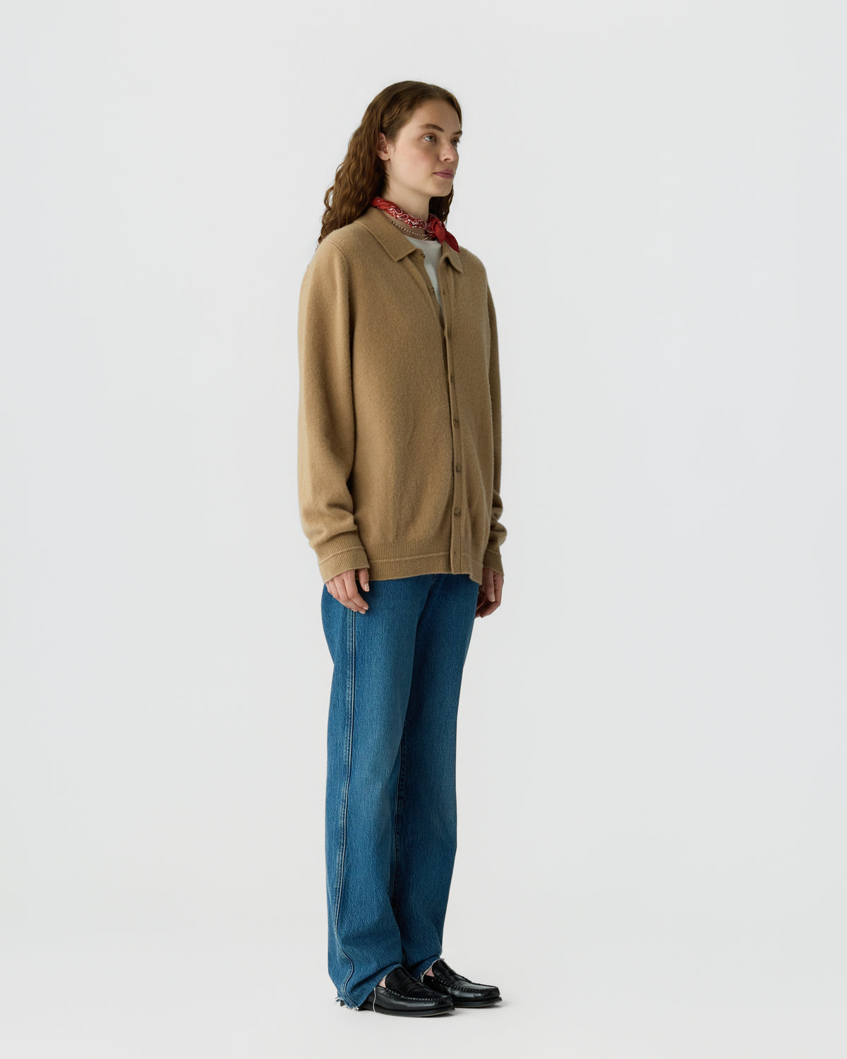 Woman wearing a light brown button-up cashmere cardigan over a white top, paired with blue jeans, black shoes, and a red patterned neck scarf, standing against a plain background.