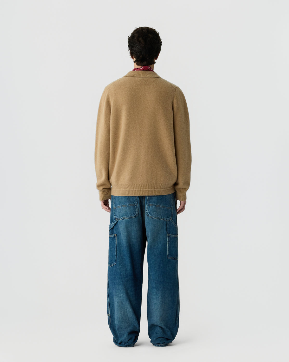 Person wearing a tan cashmere sweater and loose blue jeans standing facing away against a plain light background.