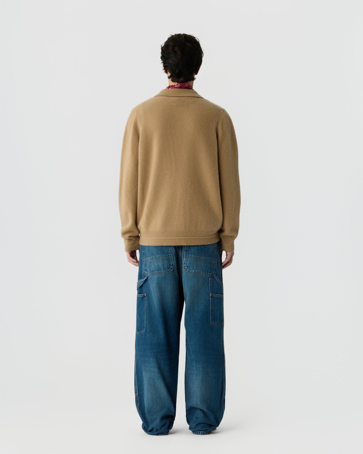 Person wearing a tan cashmere sweater and loose blue jeans standing facing away against a plain light background.