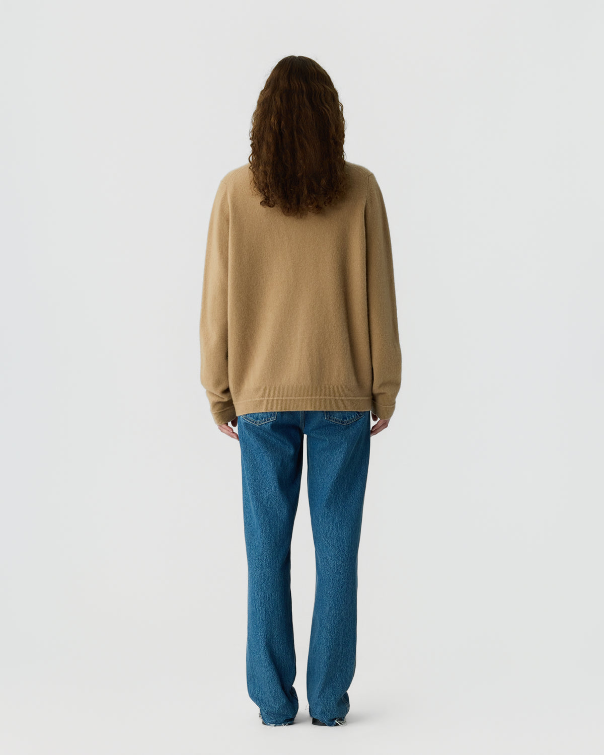 Person with long curly hair wearing a beige cashmere sweater and blue jeans standing facing away against a plain light background.