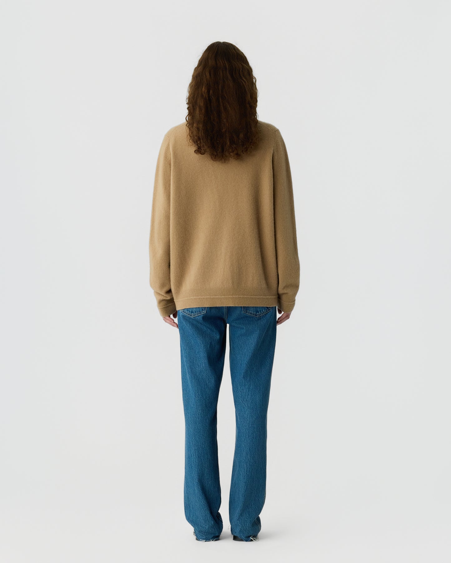 Person with long curly hair wearing a beige cashmere sweater and blue jeans standing facing away against a plain light background.