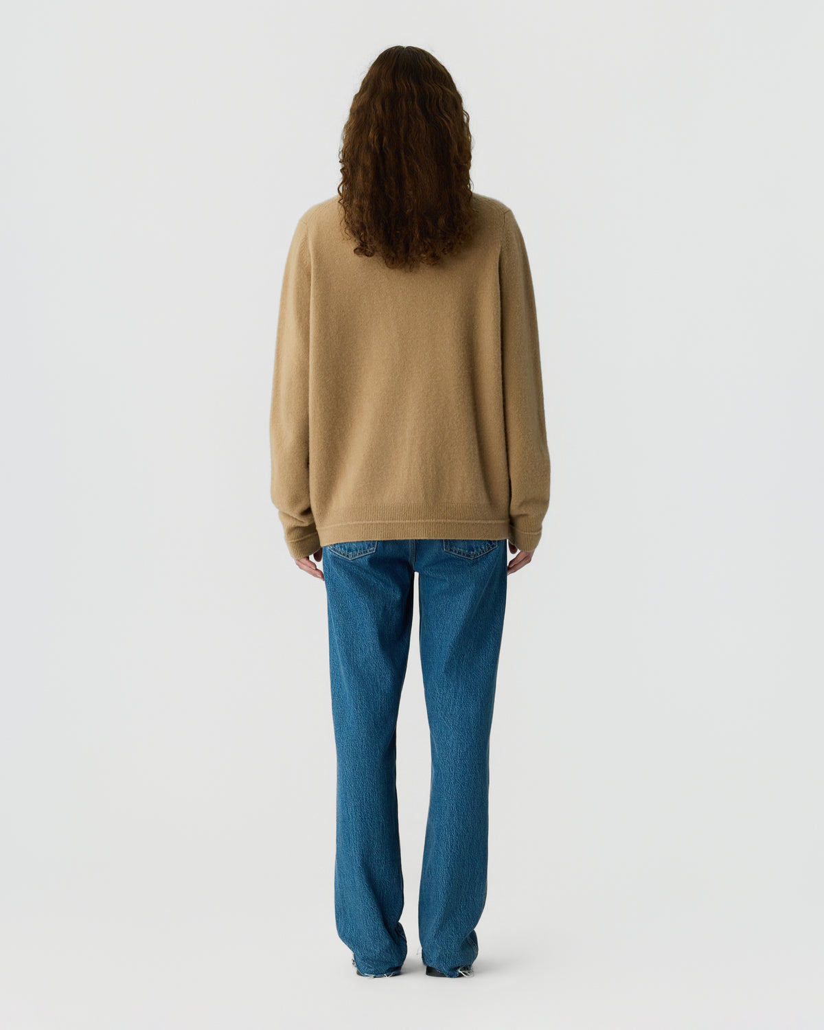 Rear view of a person wearing a loose beige sweater and blue jeans with long wavy hair against a light gray background