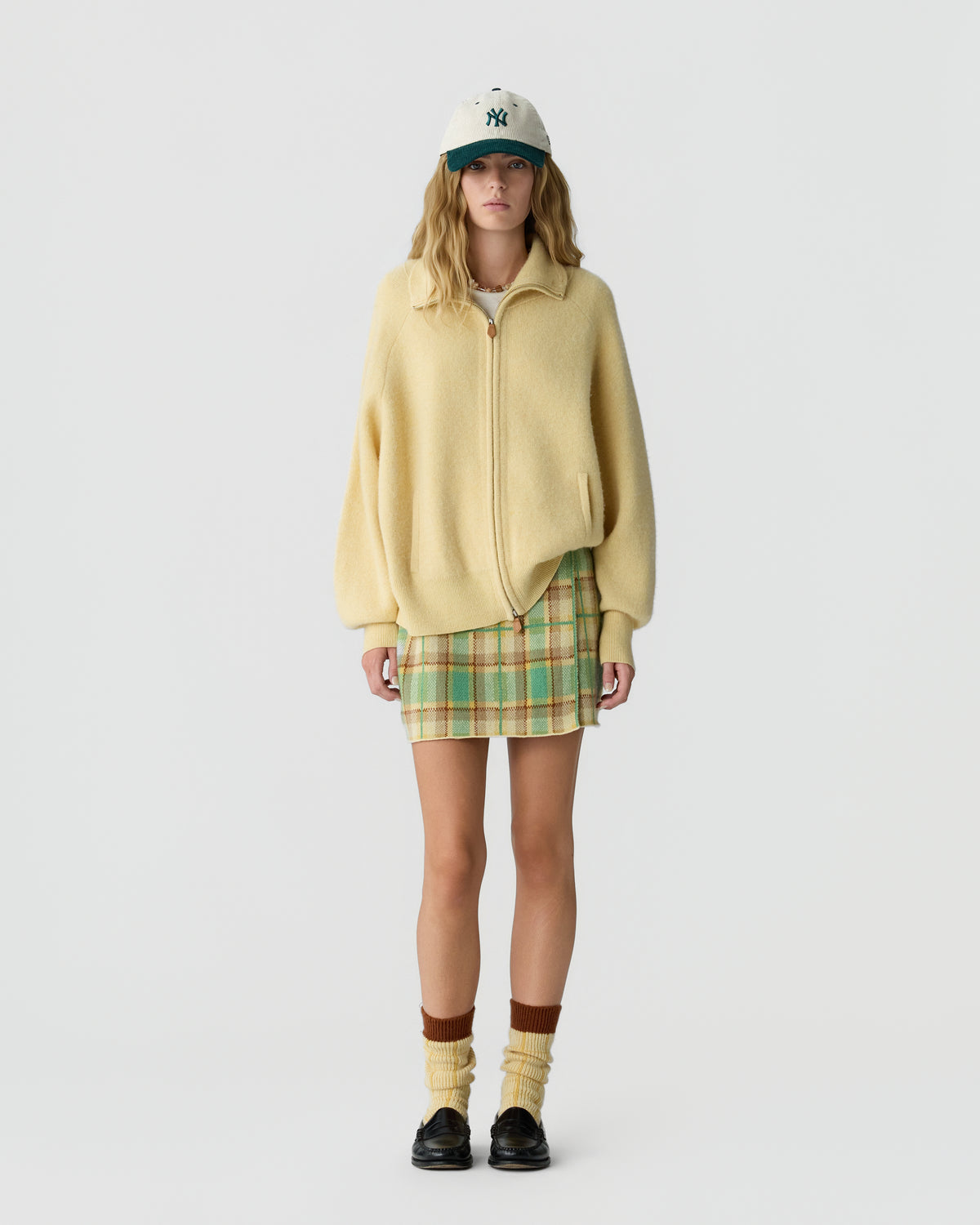 Woman wearing a pale yellow zip jacket with a plaid mini skirt and baseball cap paired with cream socks and black loafers against a light gray background