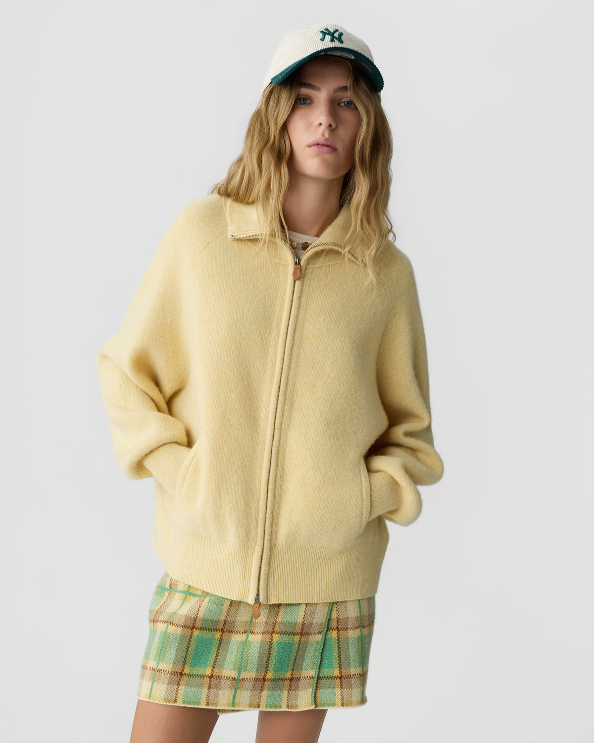 Woman wearing a pale yellow zip jacket with front pockets and a green plaid skirt while standing against a light gray background and wearing a dark baseball cap