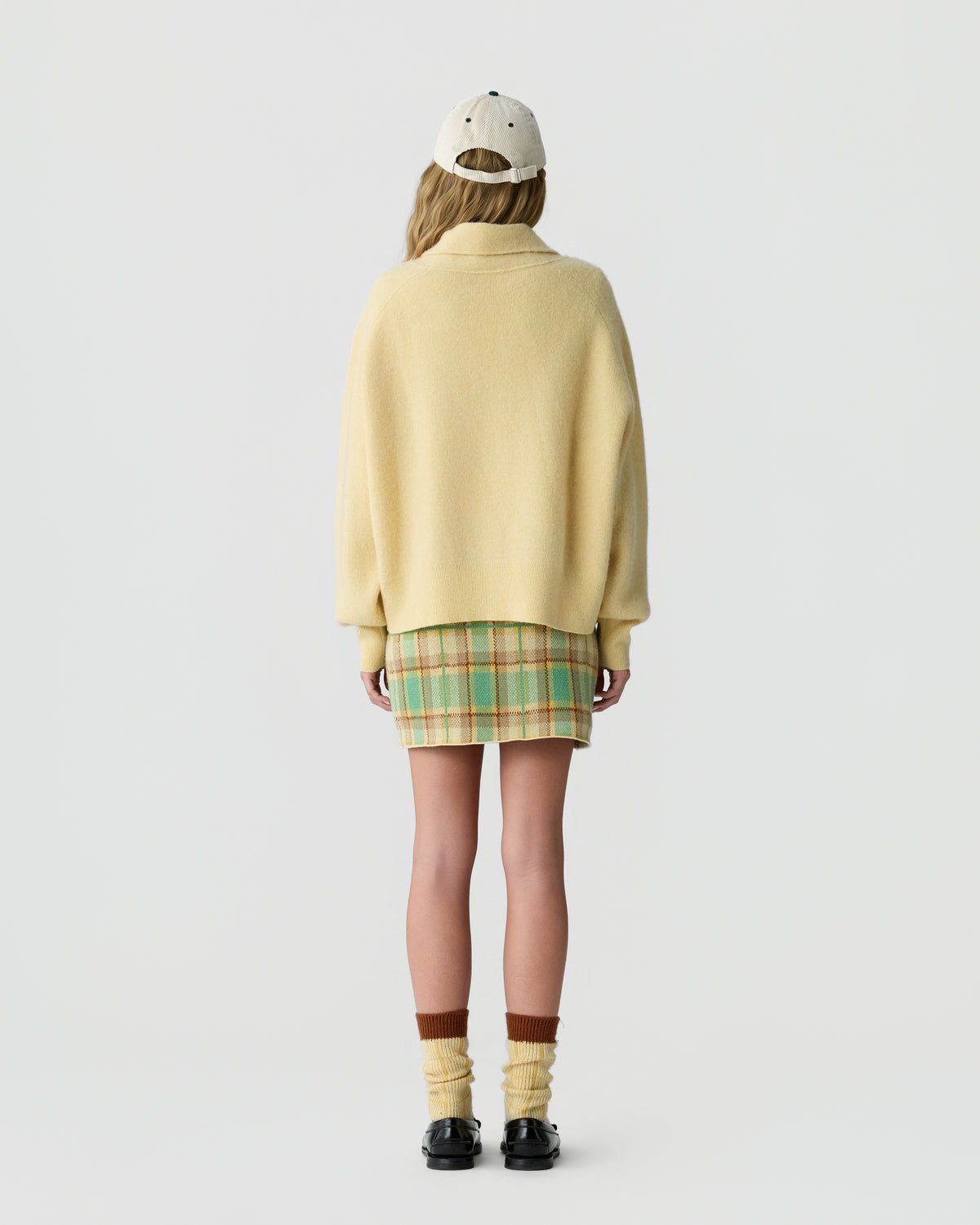 Person seen from behind wearing a pale yellow cashmere sweater with a plaid skirt a baseball cap striped socks and black loafers against a light background