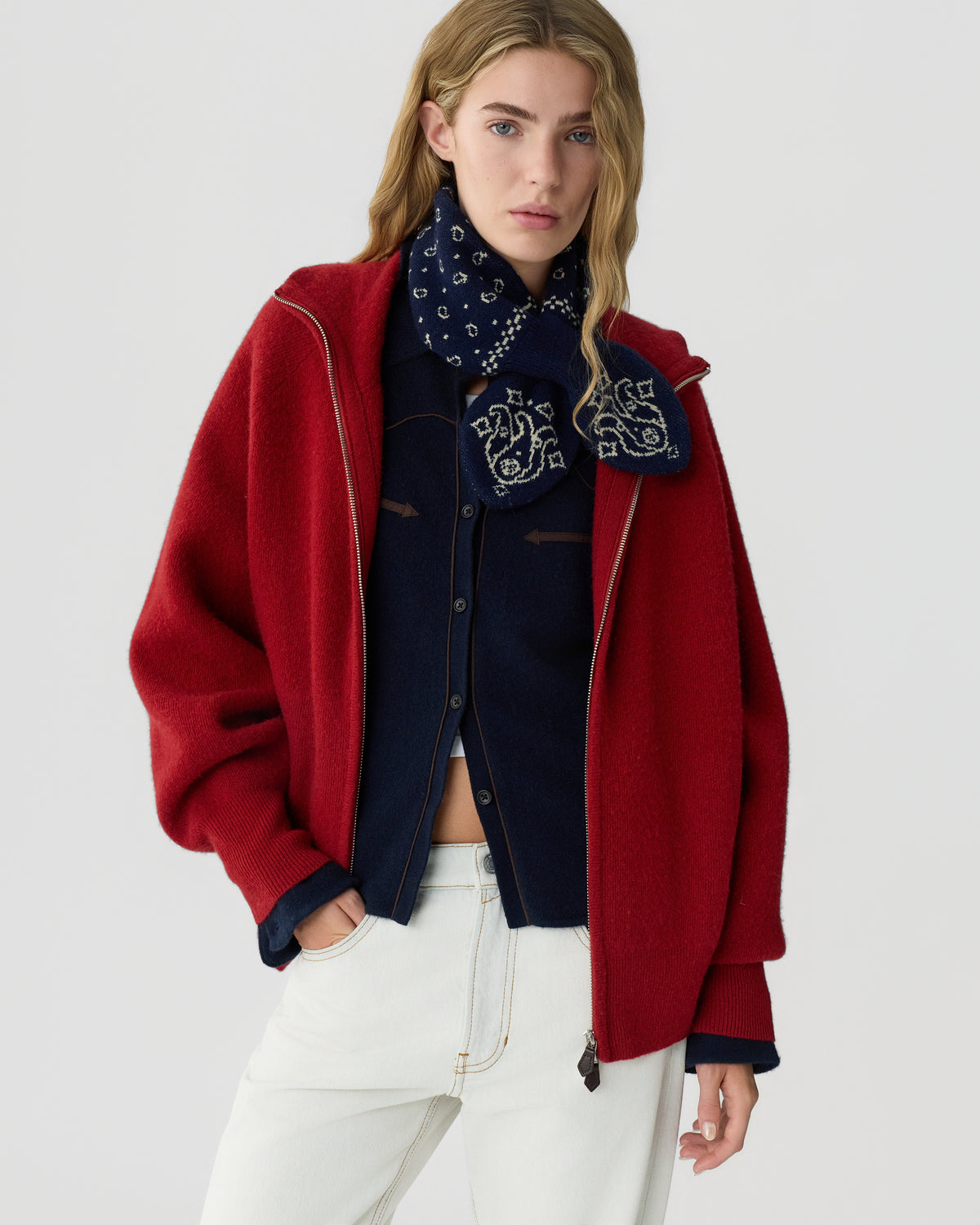 Woman wearing a red zip jacket over a navy shirt and patterned scarf, paired with cream trousers against a light gray background.