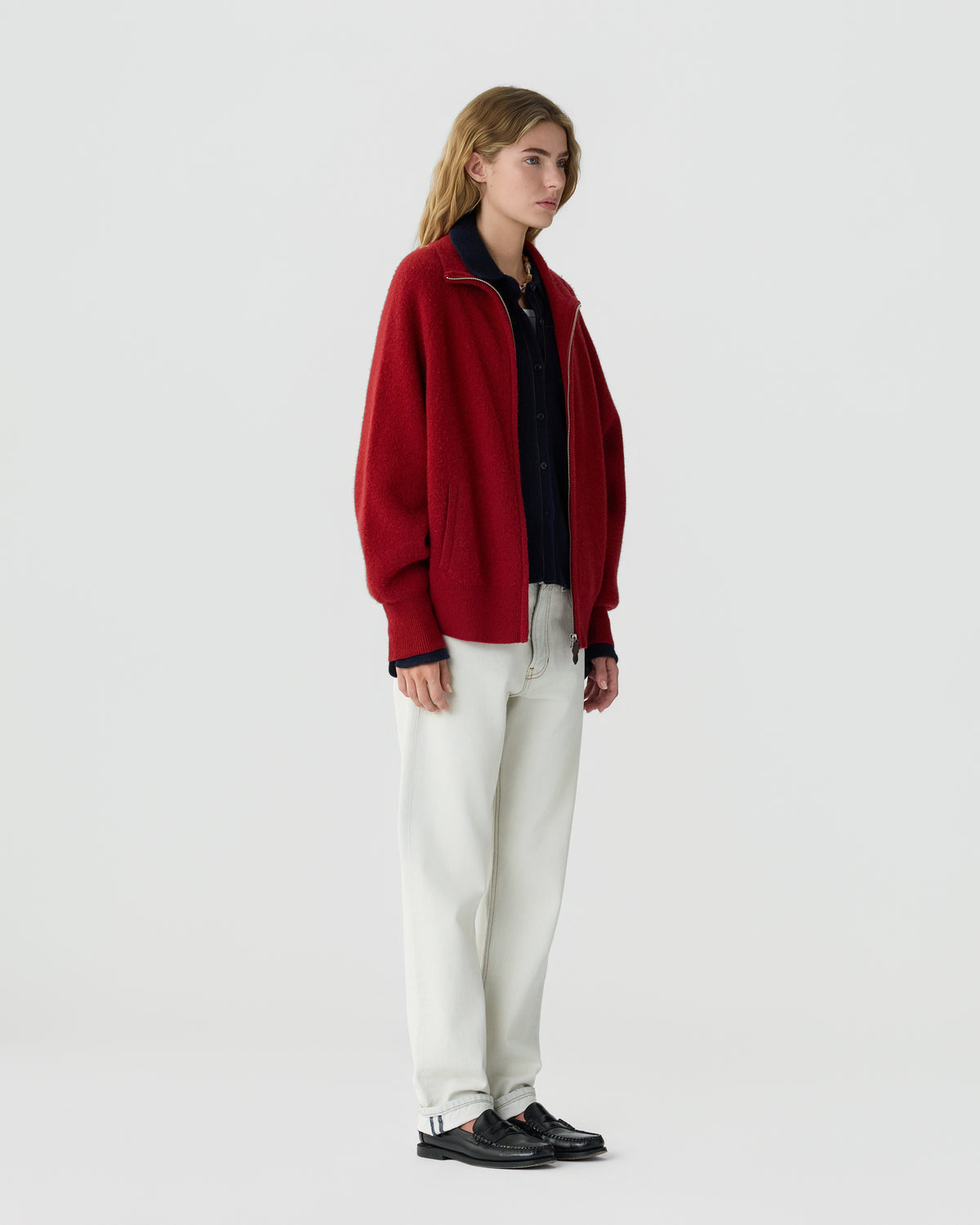 Woman standing in a red zip front sweater with a dark shirt white trousers and black shoes against a plain light gray background