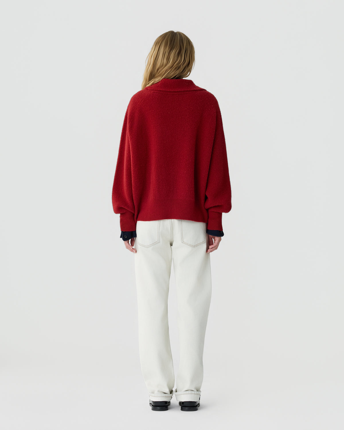 Rear view of a woman wearing a red cashmere sweater with loose sleeves white pants and black shoes against a plain light gray background
