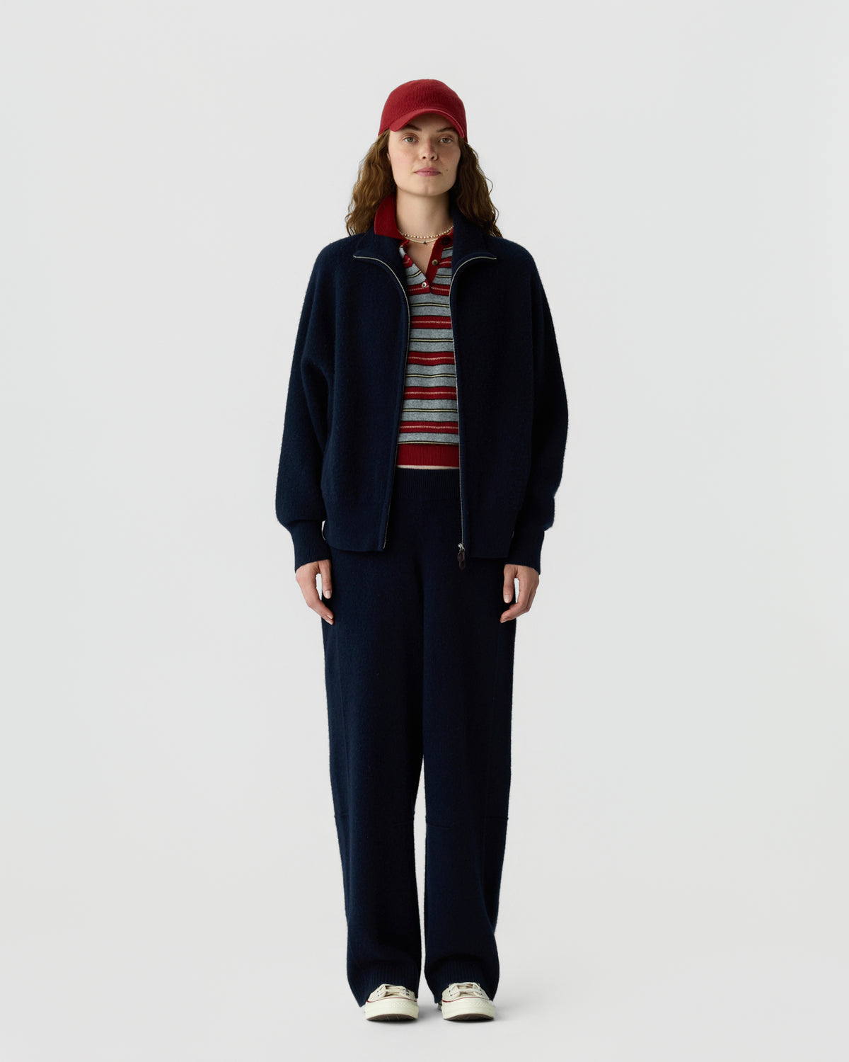 Full length person wearing a navy zip jacket and matching trousers with a striped shirt red cap and white sneakers against a light gray background