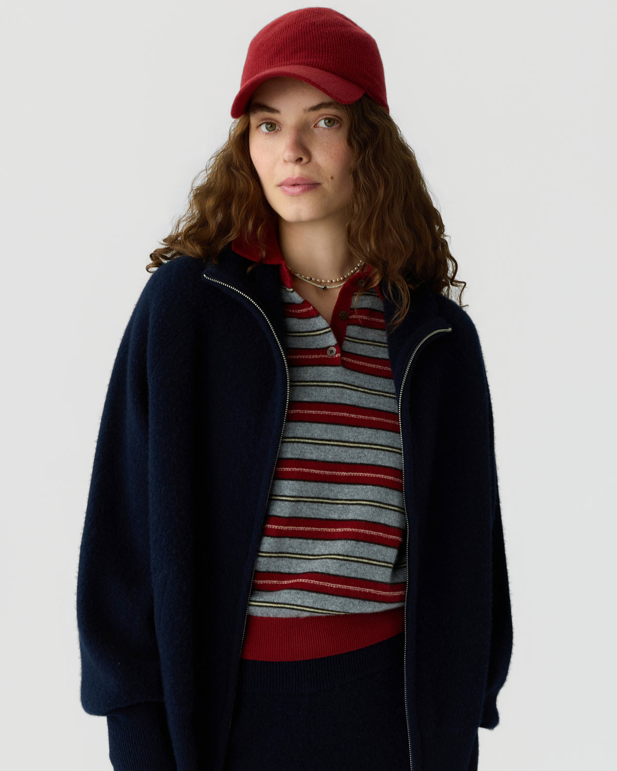 Woman with curly hair wearing a red cap and striped collared shirt under a dark zip jacket against a light gray background