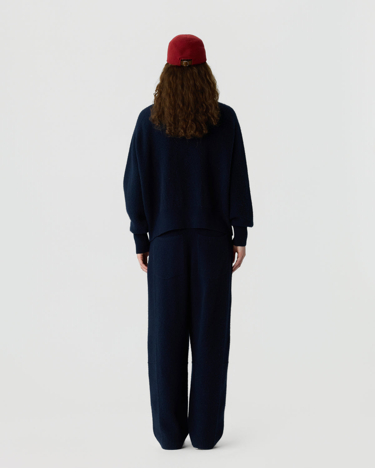 Back view of a person with long brown hair wearing a red beanie and a loose navy sweater with matching wide leg trousers against a light background