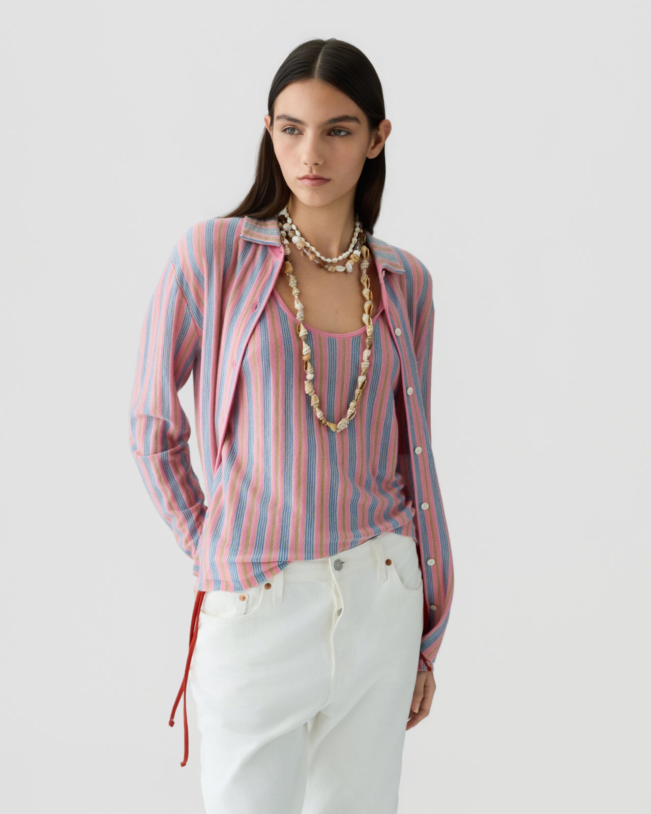 Young woman with dark hair wearing a pink and blue vertical striped lightweight shirt over a matching tank top, paired with white pants and layered shell necklaces.