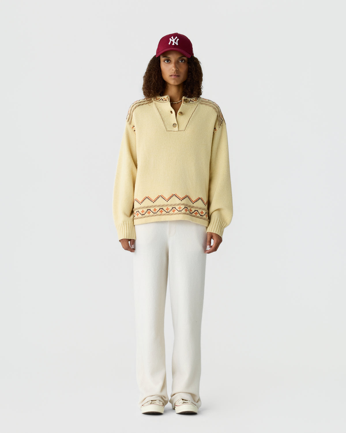 A woman wearing a cream cashmere pullover with patterned trim, white trousers, sneakers, and a burgundy baseball cap against a light background