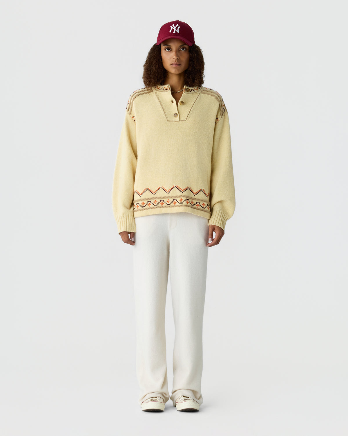Model wearing a pale yellow knit sweater with a patterned hem, white pants, cream sneakers, and a burgundy baseball cap against a light gray background.