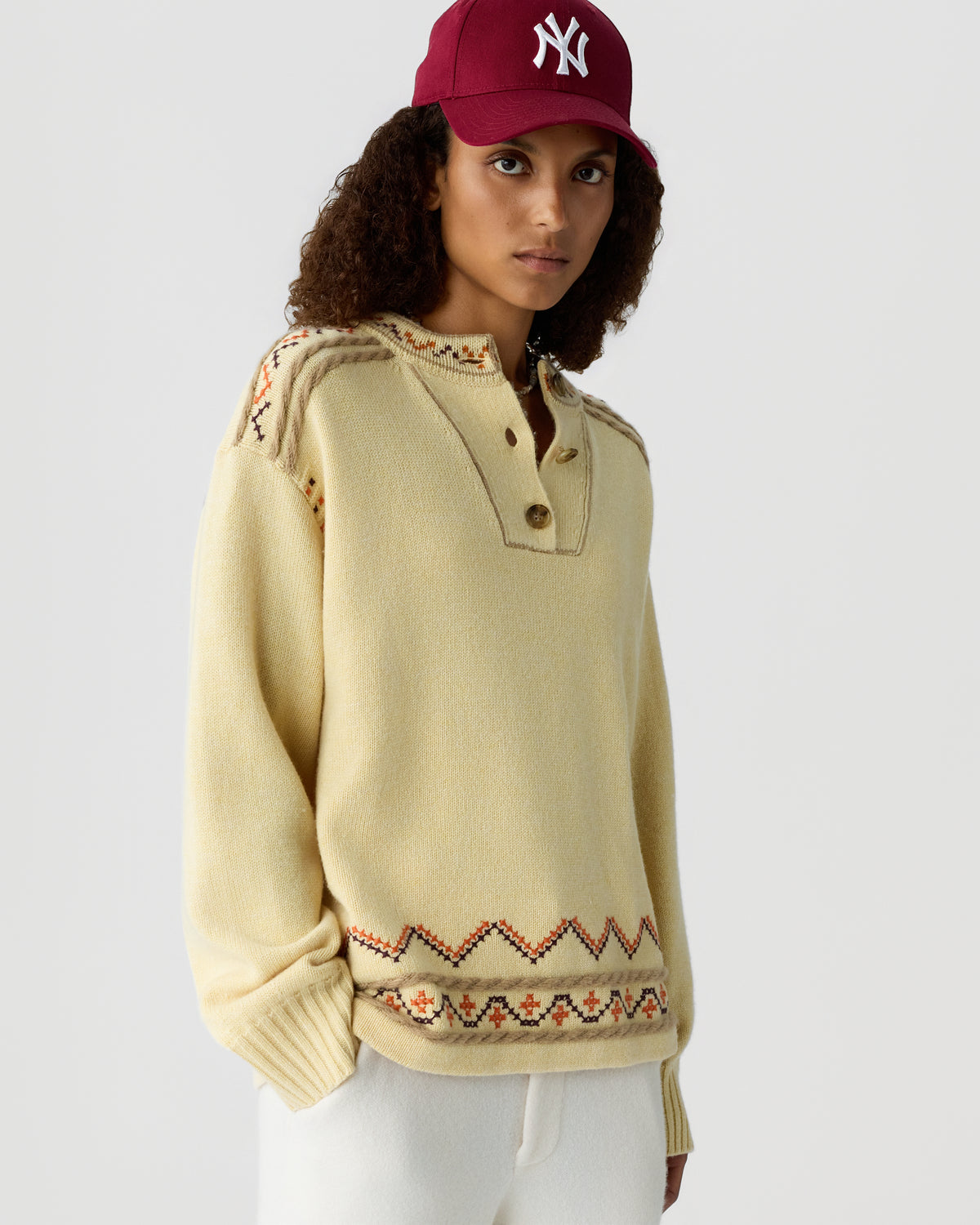 Model wearing a cream patterned knit sweater with a buttoned collar and burgundy baseball cap featuring an NY logo, posed against a light background