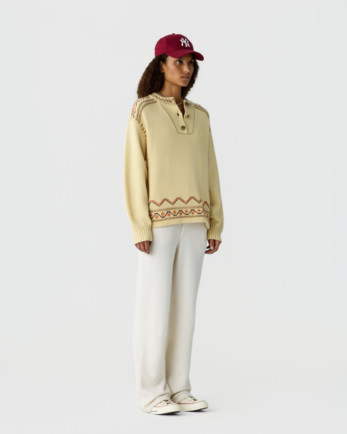 Model wearing a cream knit sweater with patterned hem, white trousers, white sneakers, and a burgundy baseball cap against a light gray background