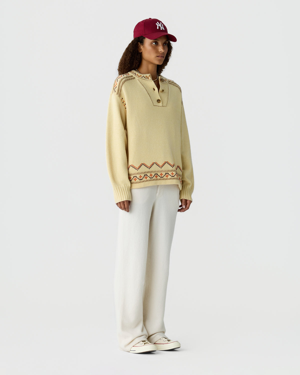 Model wearing a cream knit pullover with patterned trim and white wide leg trousers with sneakers and a burgundy baseball cap.
