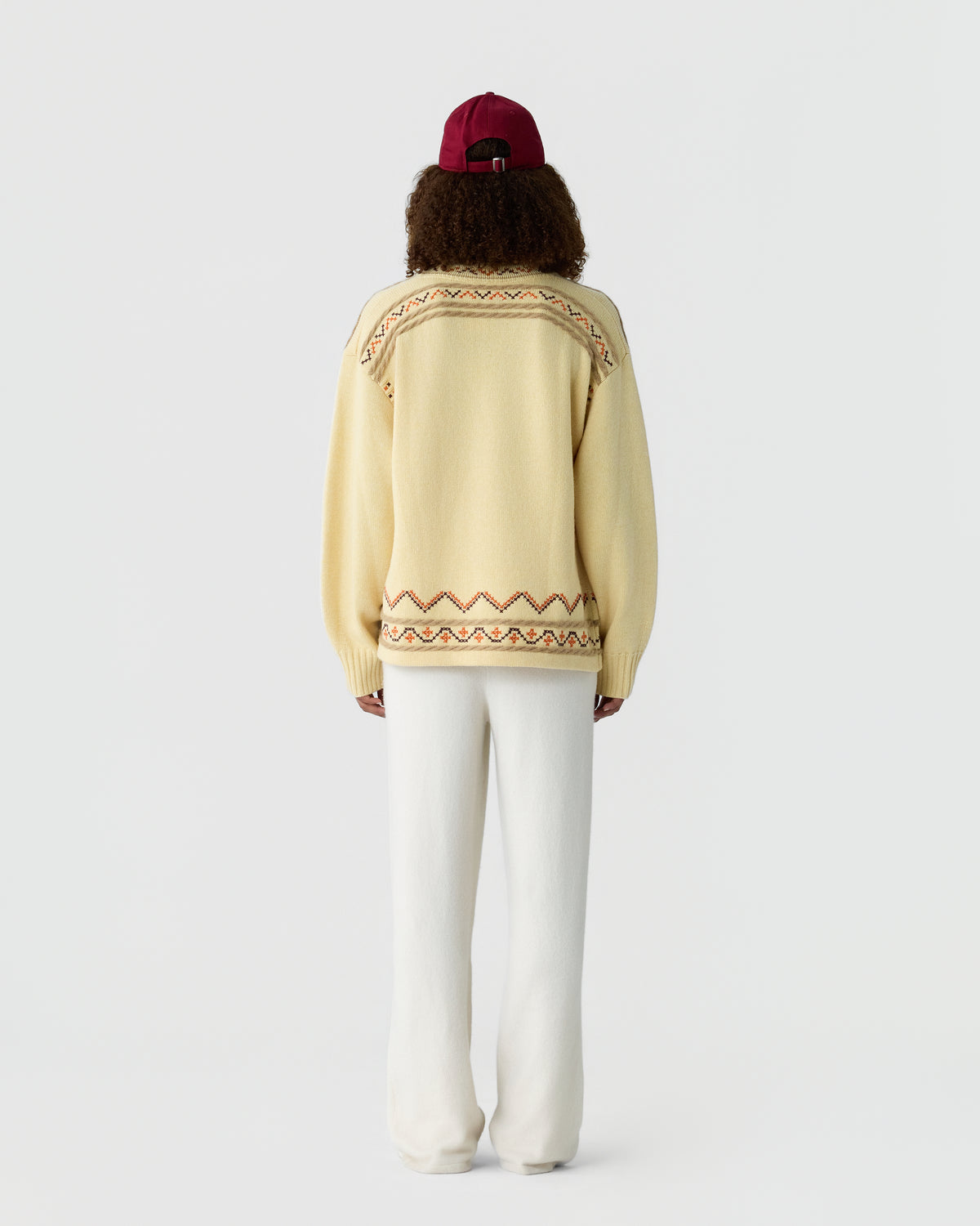 Back view of a person wearing a cream knit sweater with patterned trim white trousers and a burgundy cap against a light gray background